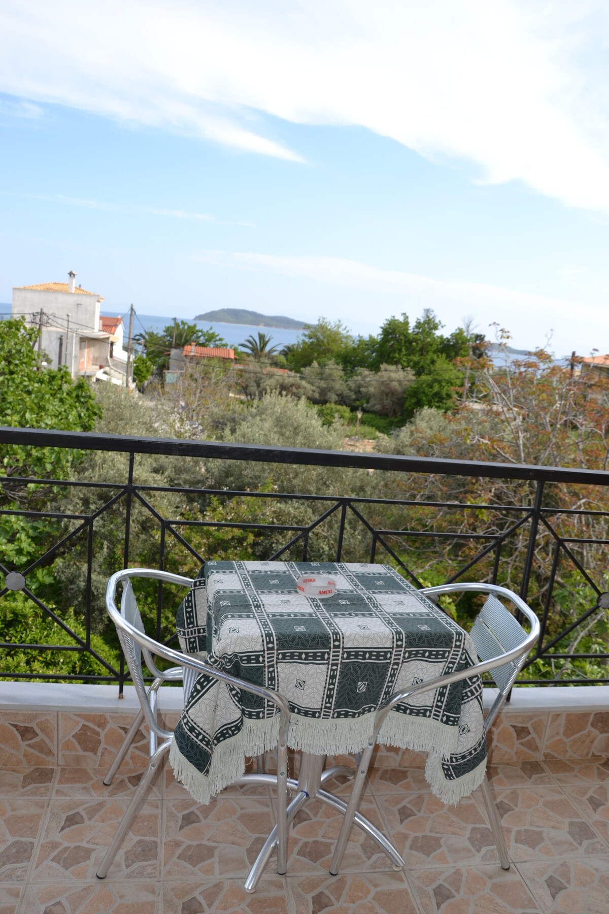 A small outdoor balcony is featured with a round table covered by a patterned tablecloth. Two metal chairs are positioned alongside, with greenery and distant hills visible in the background, providing a peaceful view of the surrounding area.
