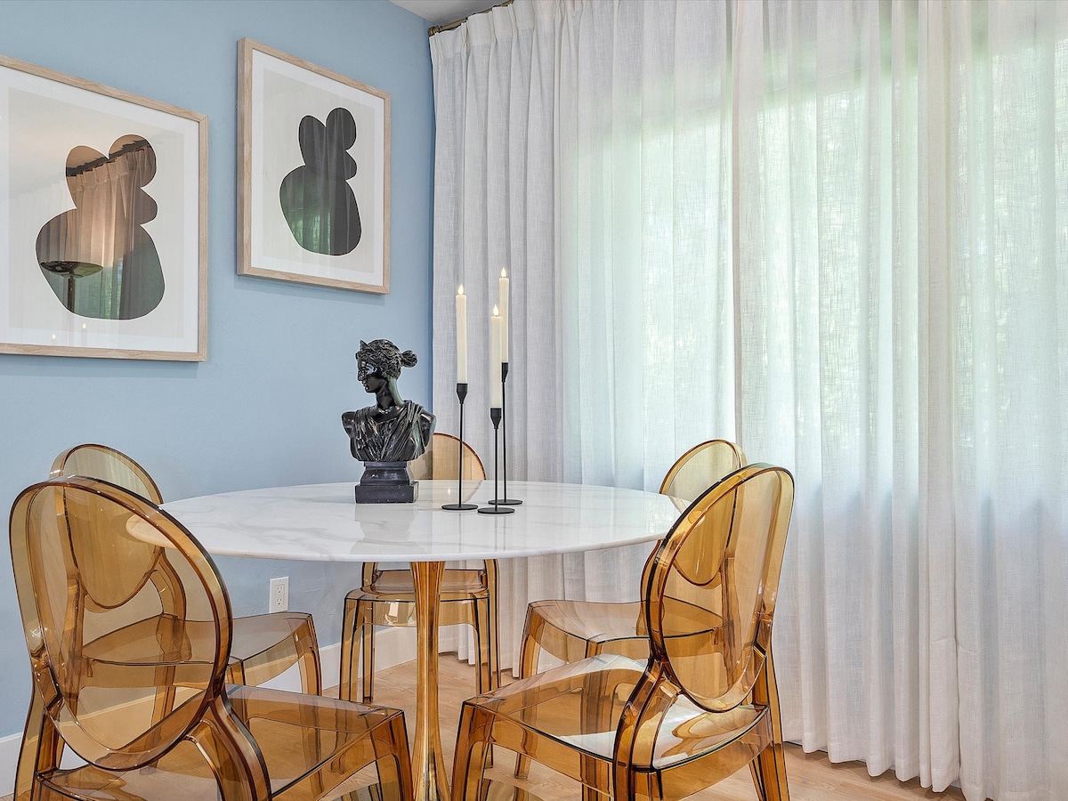 A modern dining area features a round marble table surrounded by four transparent acrylic chairs. Two abstract artworks are displayed on the wall. Soft natural light filters through sheer white curtains, creating a bright and inviting atmosphere.
