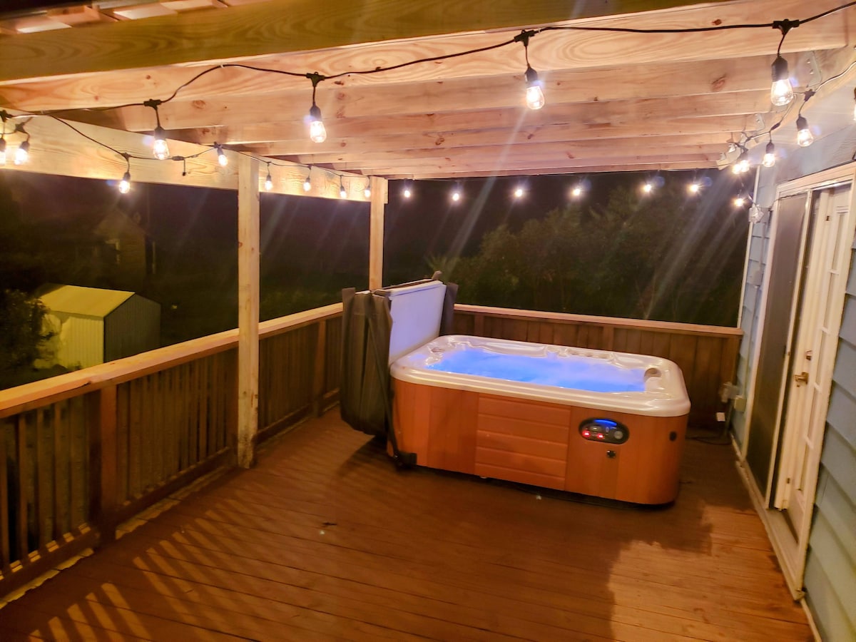 A private hot tub is positioned on a wooden deck, surrounded by string lights that cast a soft glow. The deck offers an unobstructed view of the nearby landscape, and a wooden railing enhances the outdoor experience. The entrance to the cottage is visible in the background.