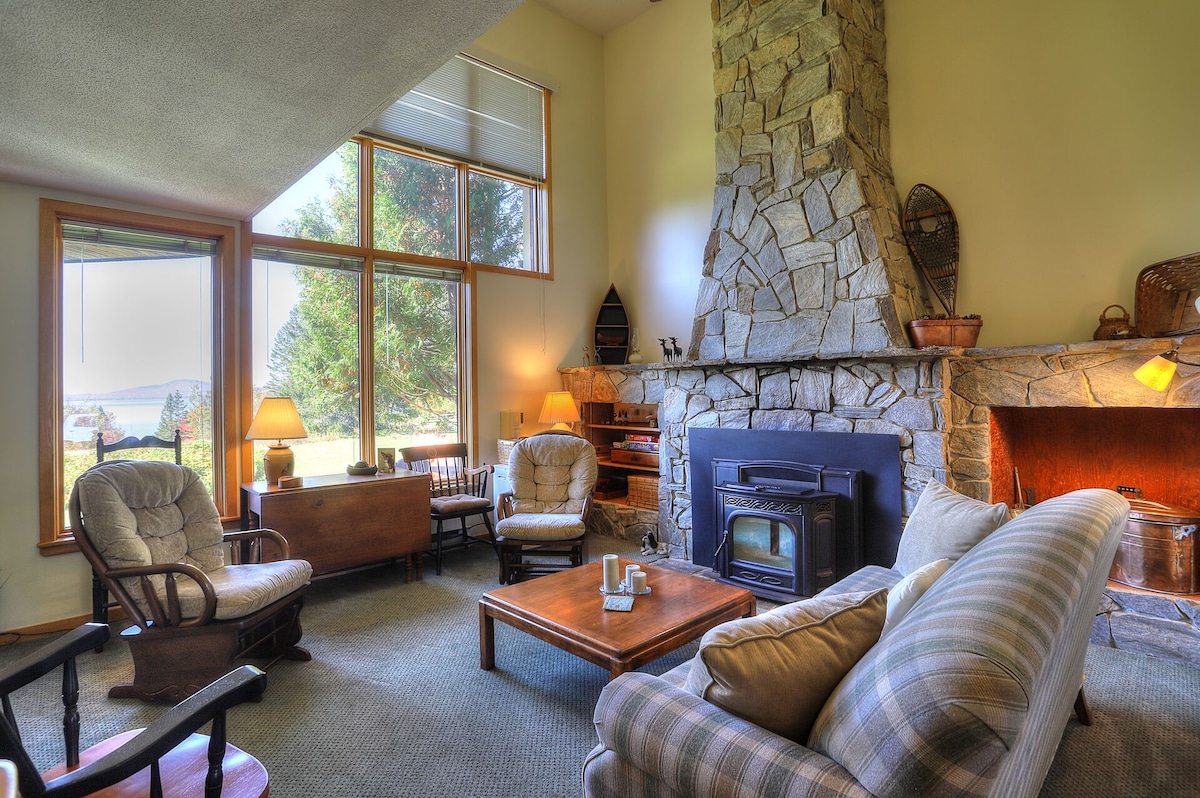 A cozy living room is presented with a stone fireplace as the focal point. Comfortable seating includes two armchairs and a sofa, arranged around a wooden coffee table. Large windows allow natural light and provide a view of the surrounding landscape.