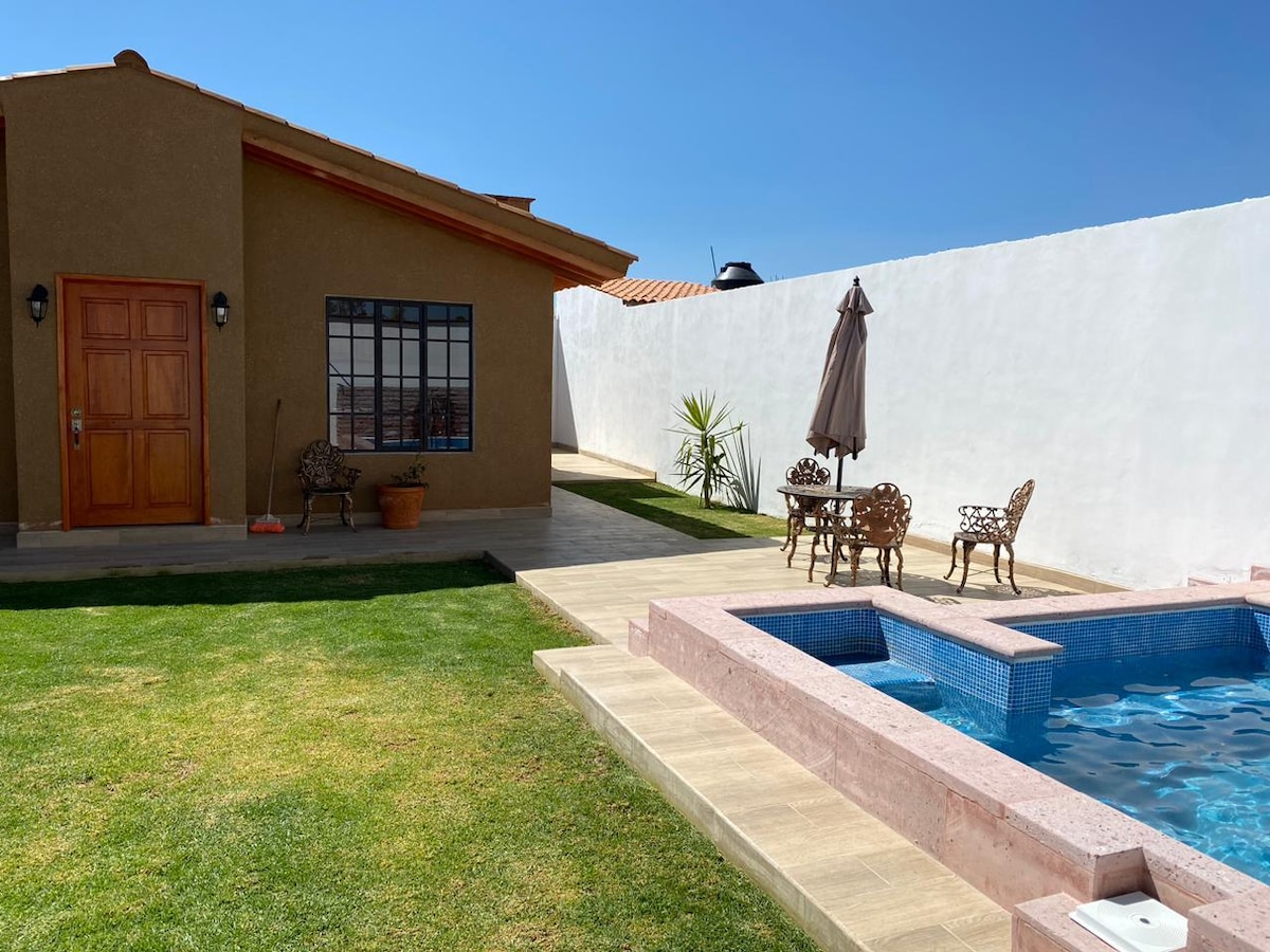 A spacious outdoor area features a well-maintained lawn, complemented by a hot tub and a small pool. A shaded table with four chairs is present, alongside a tall umbrella. The house stands nearby, showcasing a wooden door and a large window that allows natural light to flow in.