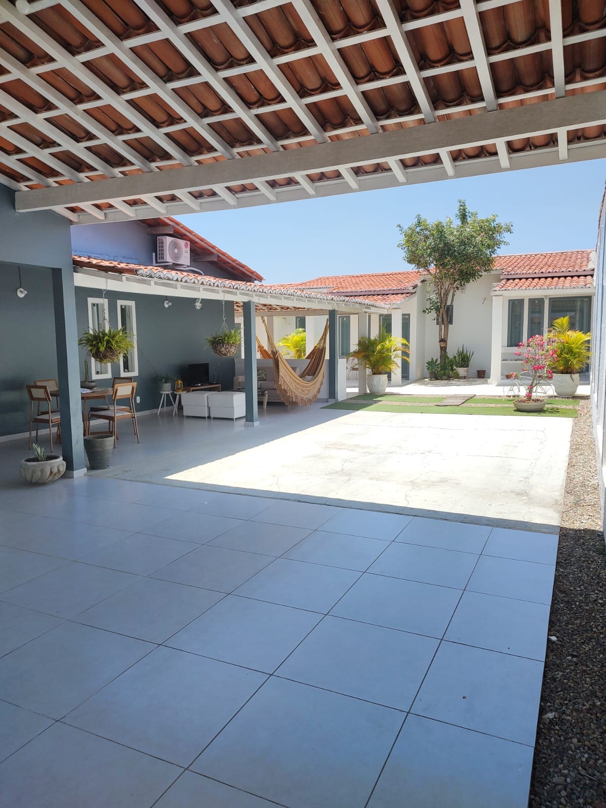 A spacious outdoor area is presented, featuring tiled flooring and well-maintained greenery. Comfortable seating is arranged on one side, with a hammock hanging between two structures. Sunlight brightens the space, highlighting the vibrant flowers and several windows that provide natural light to the nearby rooms.