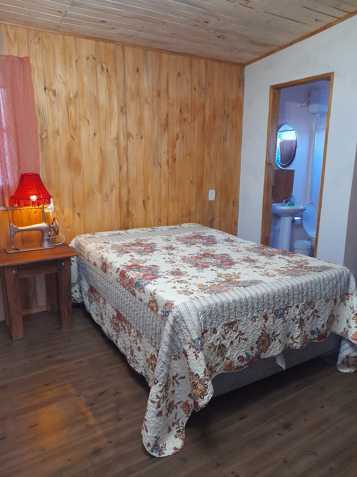 A cozy bedroom is presented with a wooden wall finish and warm lighting. A spacious bed is covered with a floral patterned quilt. A bedside table with a lamp is situated next to the bed, and a bathroom entrance is visible in the background.