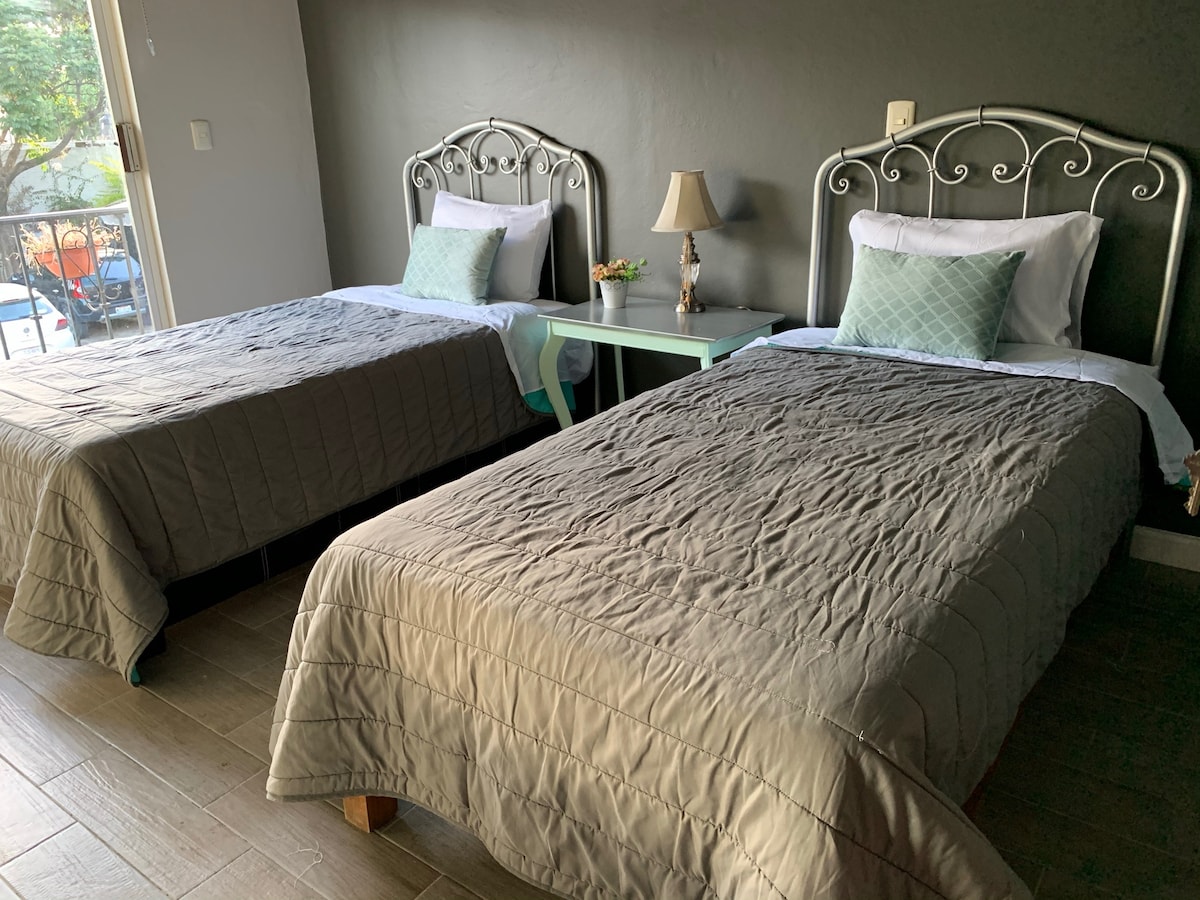 The room features two spacious beds, each with neutral-colored bedding. A small lamp sits on a bedside table between them, adorned with a potted plant. Natural light filters in from a glass door that leads outside.