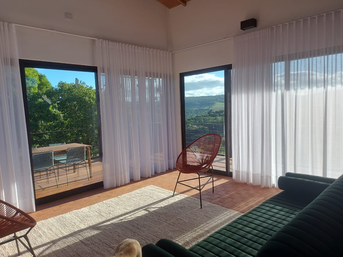 A bright living space features large glass doors providing a view of the outdoor seating area with a dining table. Lightly draped sheer curtains add softness to the room, which is furnished with a dark green sofa and a textured area rug.