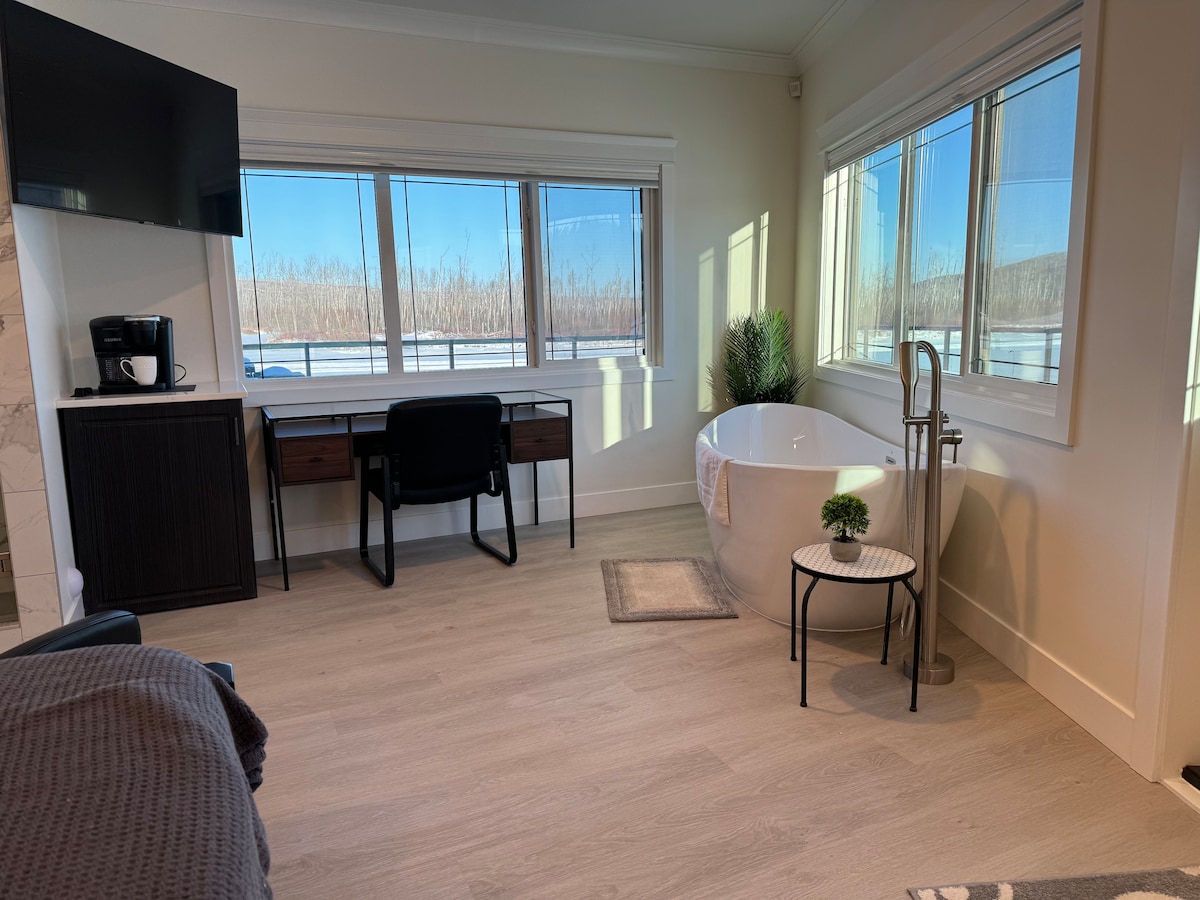A bright and airy bathroom area features a freestanding bathtub beside a small table with a plant. A desk and chair are positioned against the window, which allows natural light to fill the space. A television is mounted on the wall, contributing to a modern design.