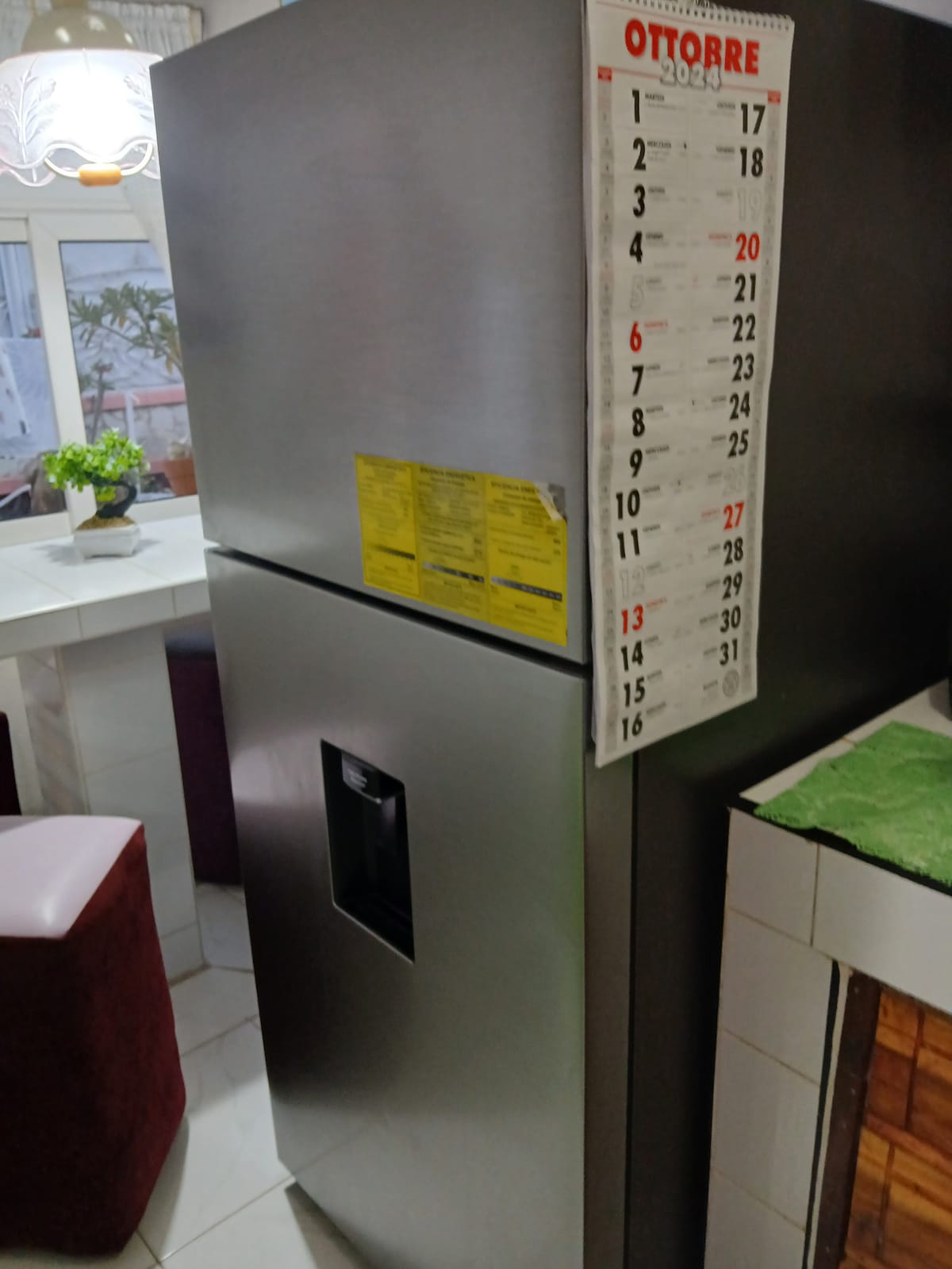 A sleek stainless steel refrigerator stands against a light-colored wall, featuring an ice and water dispenser on the front. A calendar hangs nearby, showcasing the month of October, adding a practical element to the kitchen environment.