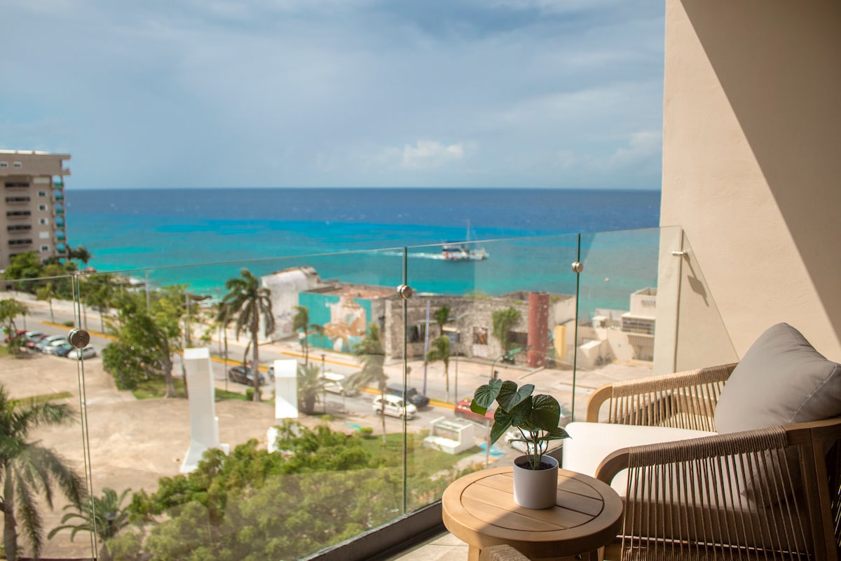 A private balcony is featured, showcasing panoramic views of the Caribbean sea. A small potted plant sits on a wooden table next to a comfortable seating area, with gentle waves and a clear sky in the background. The vibrant colors of the ocean create a serene atmosphere.