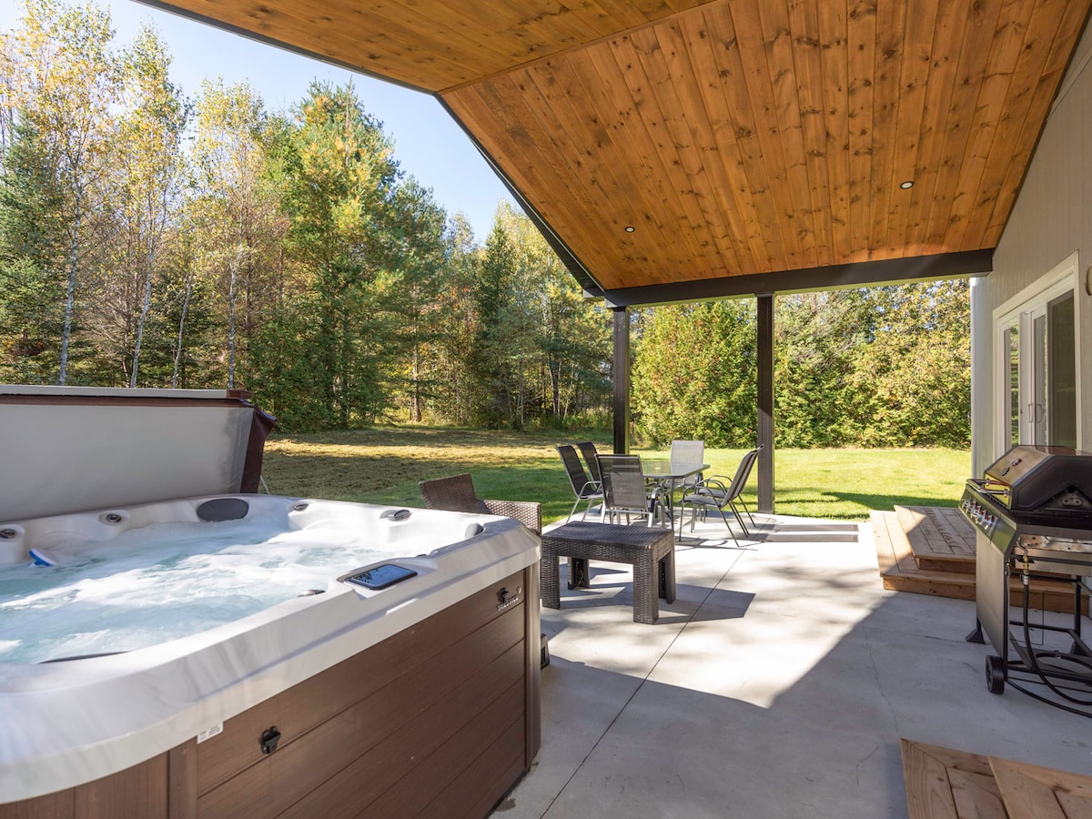 A hot tub is positioned on a shaded patio under a wooden ceiling, surrounded by trees in the background. A seating area includes metal chairs and a table, while a barbecue grill is situated nearby. Natural light filters through the outdoor space.