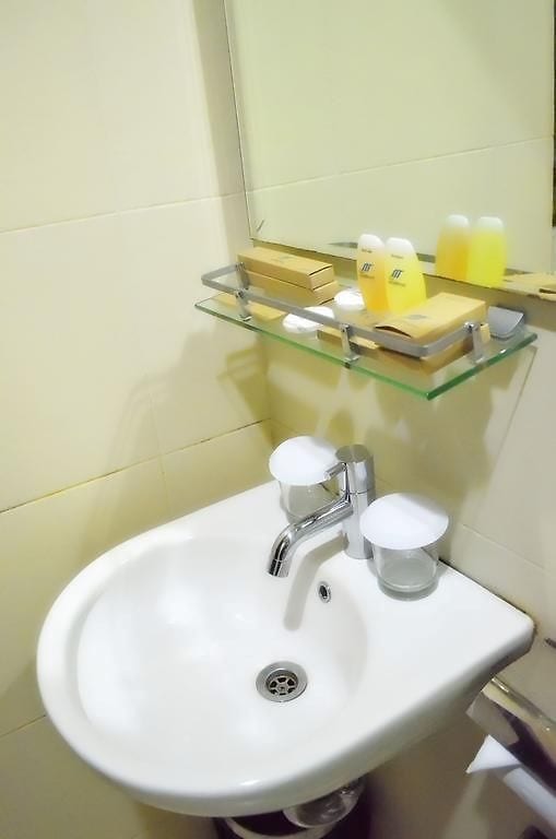 A compact bathroom sink area is presented, featuring a circular basin with a modern faucet. Shelving above holds complimentary toiletries, including small bottles and soap. Two clear glasses are positioned beside the sink, and a mirror is mounted on the wall, reflecting the minimalist design.
