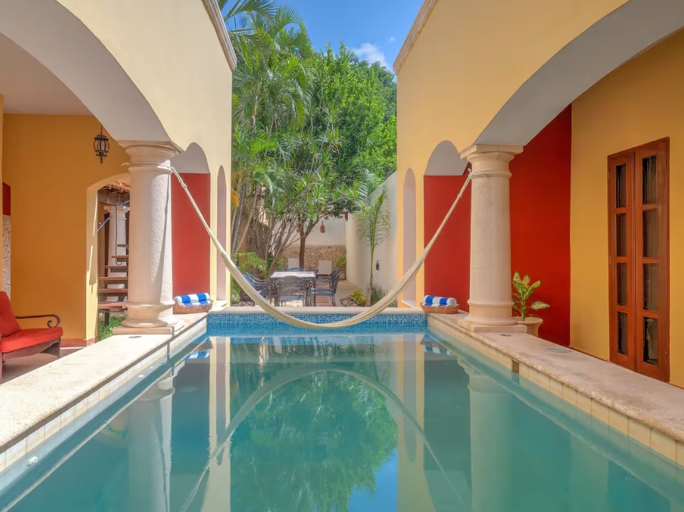 A tranquil courtyard is revealed, featuring a refreshing pool reflecting the clear sky. Pillars frame the entrance, leading to a seating area surrounded by lush greenery. A hammock is suspended between two pillars, inviting relaxation in the serene environment.
