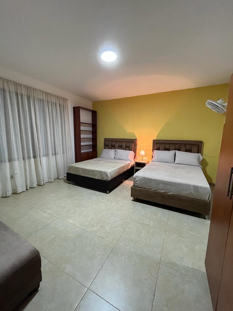 A well-lit room features two spacious beds with upholstered headboards. A sectional sofa is positioned nearby. Light curtains frame a large window, allowing soft light to fill the space. A shelving unit and a wooden wardrobe are also visible against the walls.