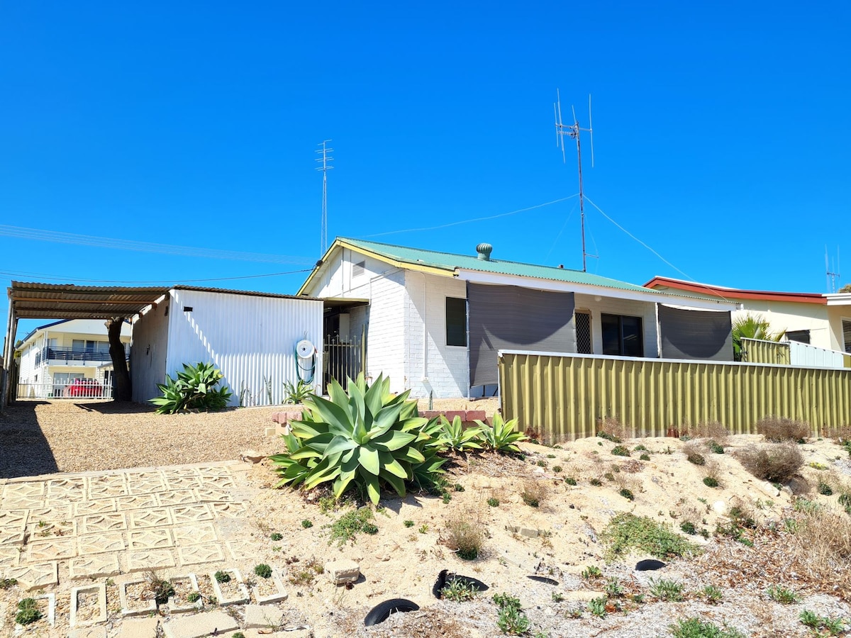 A single-story coastal cottage is visible, featuring a light-colored exterior and a green roof. Native plants and gravel landscaping surround the property. An undercover carport is present, with an adjoining area offering additional parking space. The clear blue sky adds to the serene setting.