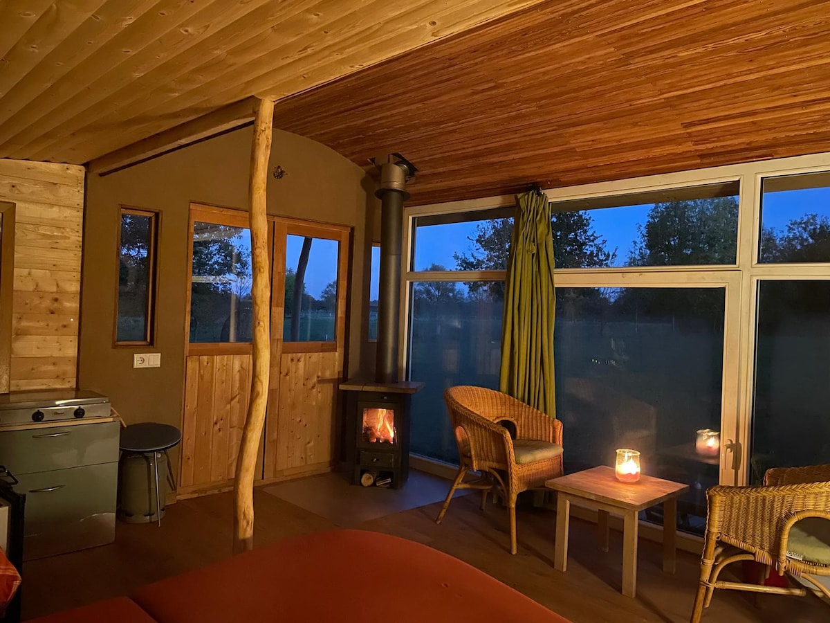 An inviting interior features warm wood paneling and a cozy wood stove. Large windows provide a view of the surrounding nature, while two rattan chairs and a small table are positioned for relaxation. A flickering candle adds a soft glow to the space.