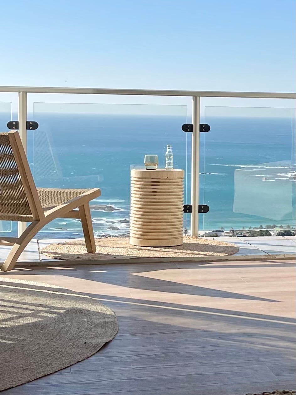 A wooden chair is placed on a balcony, accompanied by a textured side table featuring a glass and a bottle. A clear view of the Atlantic Ocean and coastline is visible in the background, enhanced by natural light illuminating the area.
