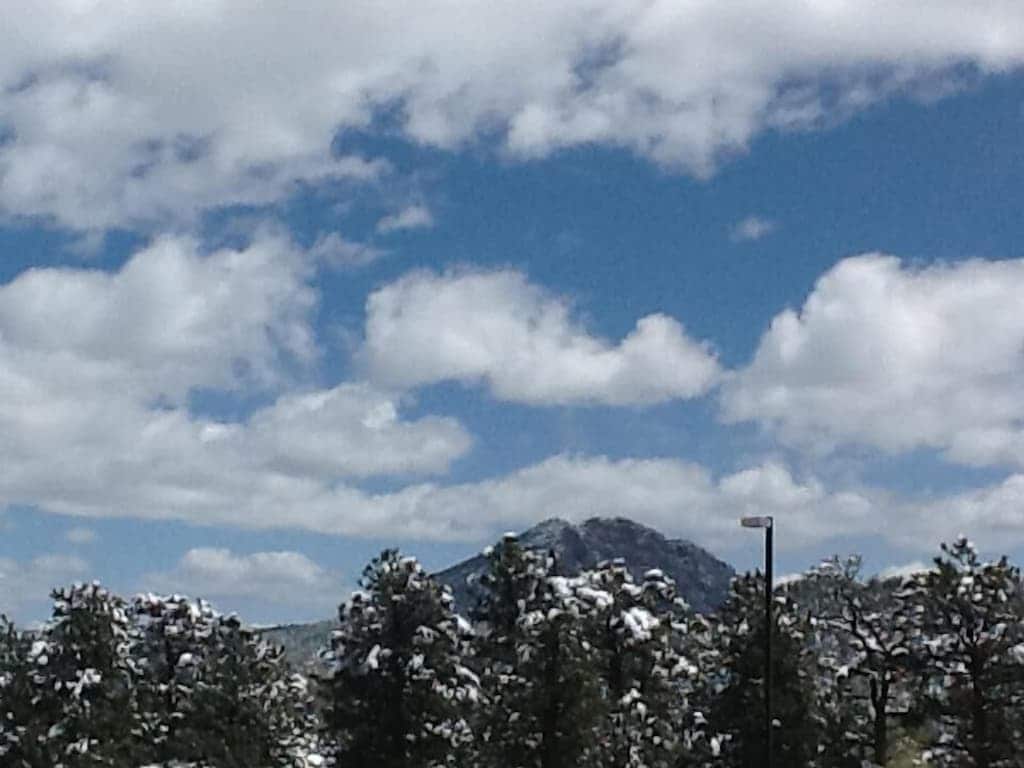 A clear blue sky filled with fluffy white clouds frames a snow-capped mountain peak in the distance. Lush green trees adorned with a dusting of snow are visible in the foreground, enhancing the serene natural landscape.
