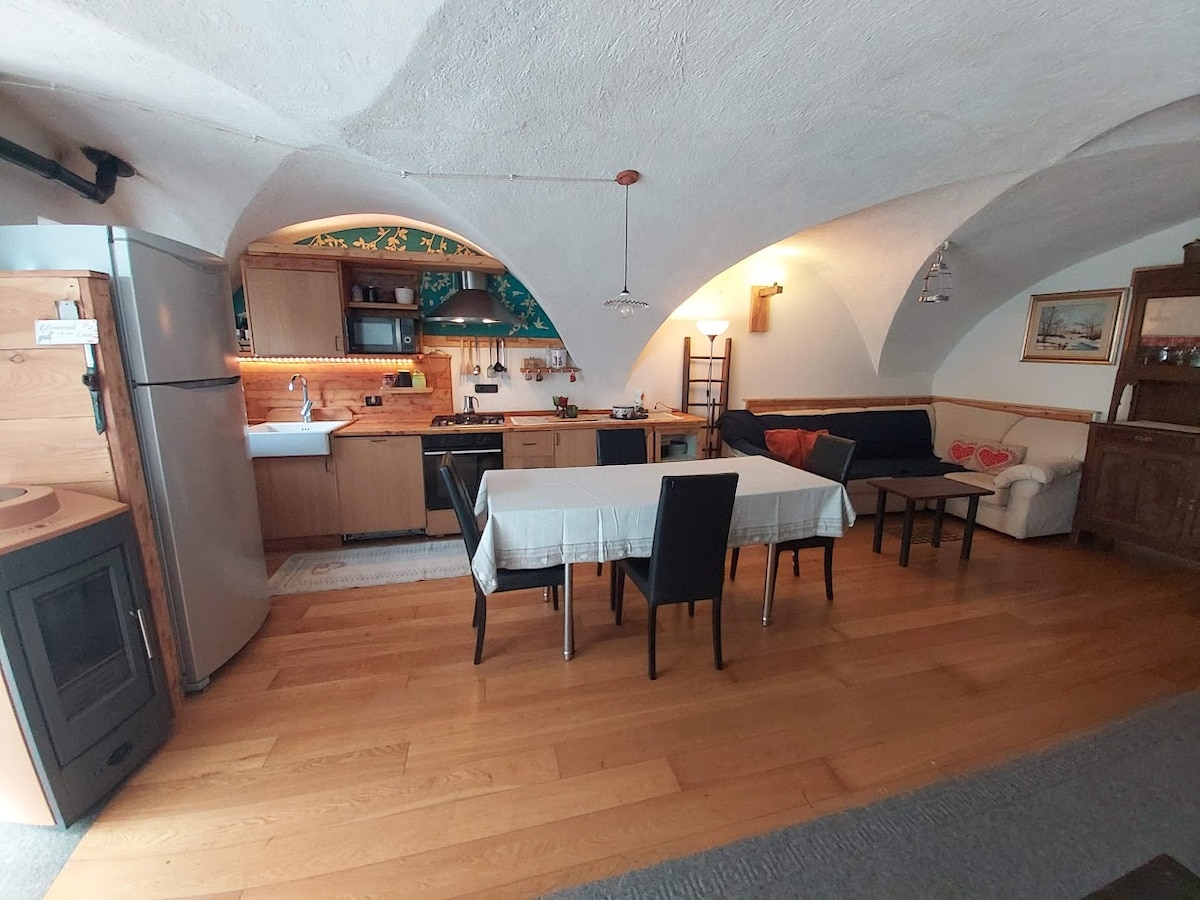An open-plan living area is presented with arched ceilings and natural wood accents. A spacious dining table with black chairs is positioned beside a well-equipped kitchen. A comfortable seating area features a sofa, while warm lighting creates a welcoming ambiance.