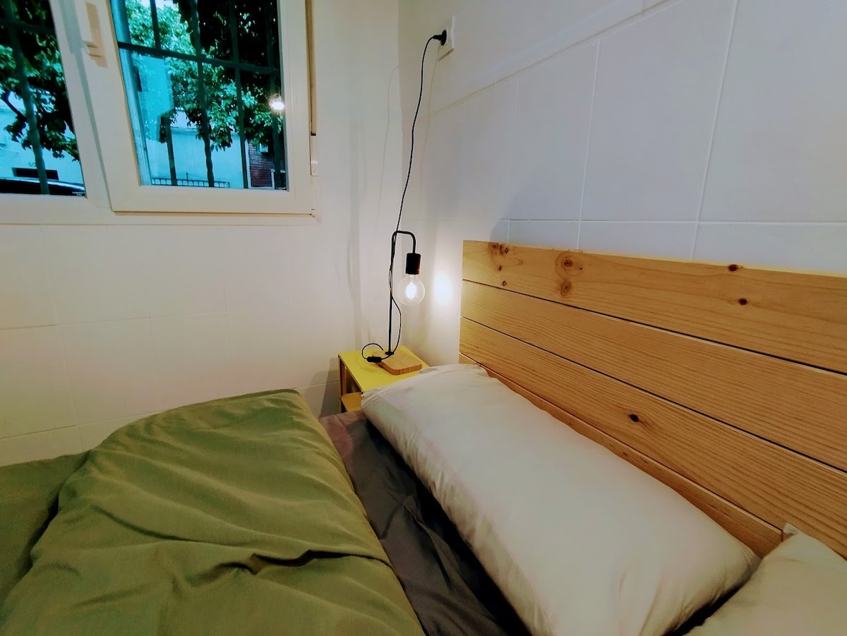 A cozy sleeping area features a wooden headboard and soft bedding in neutral tones. A small bedside table holds a book and a lamp with an exposed bulb. Natural light filters in through the nearby window, offering a calming atmosphere.