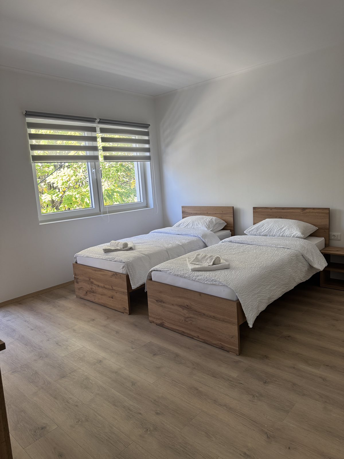 A bright bedroom features two twin beds with soft white bedding. Each bed is positioned against a wooden frame, with a window providing natural light and a view of greenery outside. Light-colored wooden flooring complements the overall design.