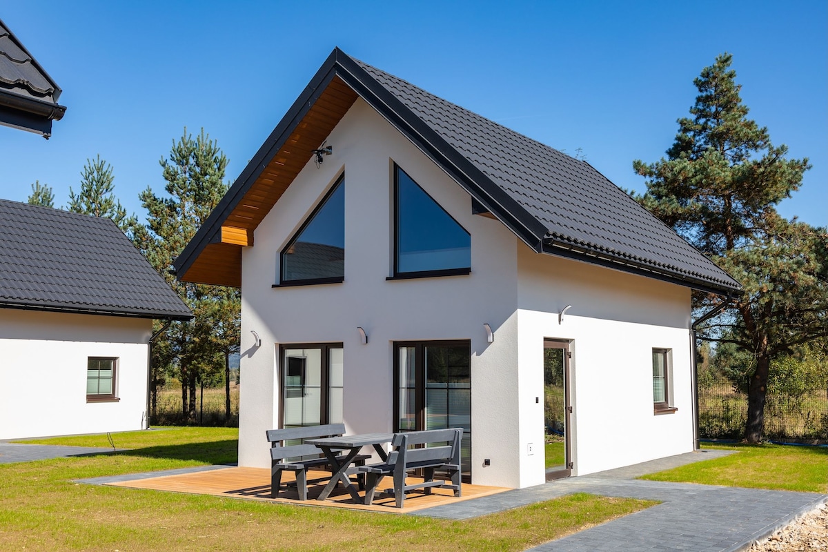 A modern two-story house is situated amidst greenery, featuring a sloped roof and expansive triangular windows that allow natural light to enter. A wooden patio with outdoor seating is visible, providing a space for relaxation surrounded by the landscaped yard.