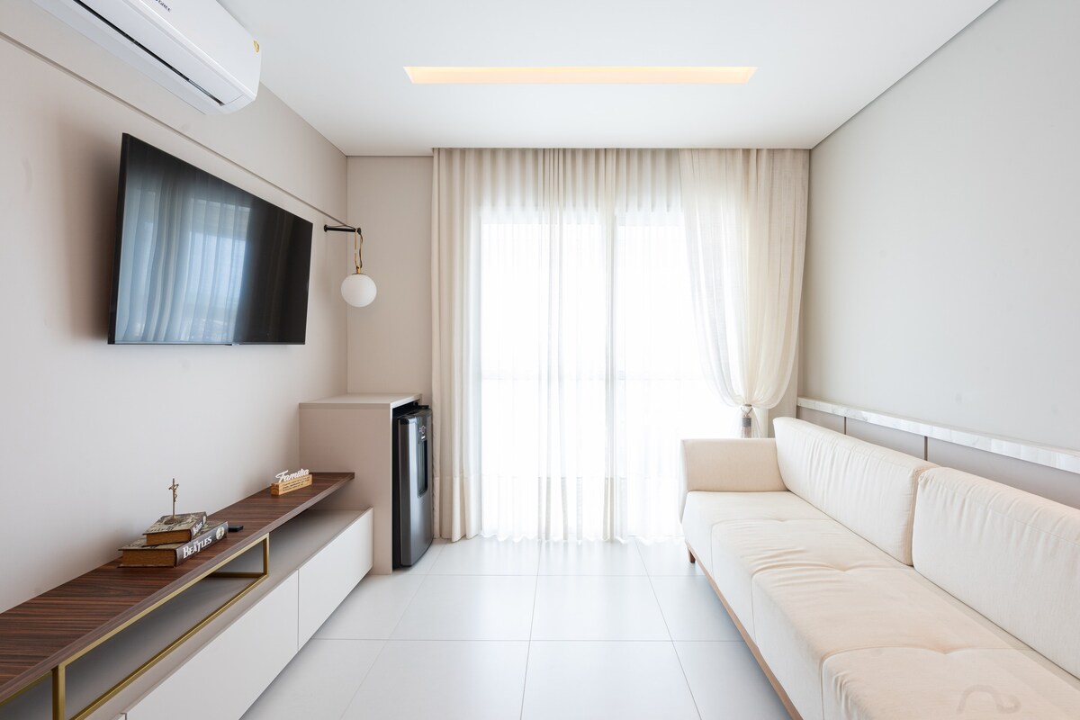 A neatly arranged living area is presented with a light-colored sofa and a modern TV mounted on the wall. A small refrigerator is visible beside a sideboard, while sheer curtains frame the window, allowing natural light to fill the space.