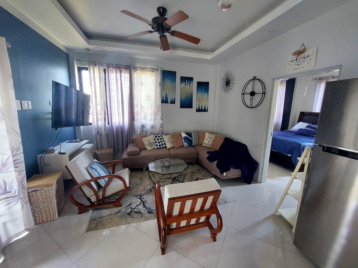 A living area is depicted with a large sectional sofa and a textured area rug. A wall-mounted television is visible, alongside light curtains that filter natural light. The space features a modern ceiling fan and a sleek refrigerator partially shown in the corner.