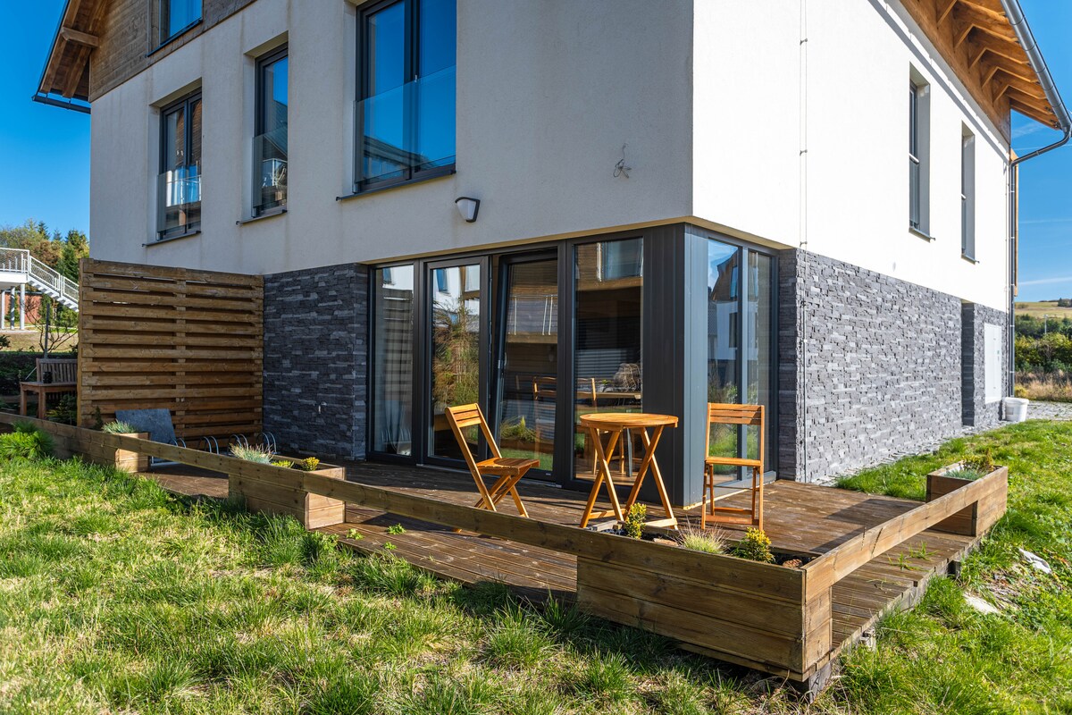 A private terrace is presented with wooden decking and includes two folding chairs and a small table. The exterior of the apartment showcases a mix of light-colored walls and stone accents, with large glass doors providing access to the outdoor space.