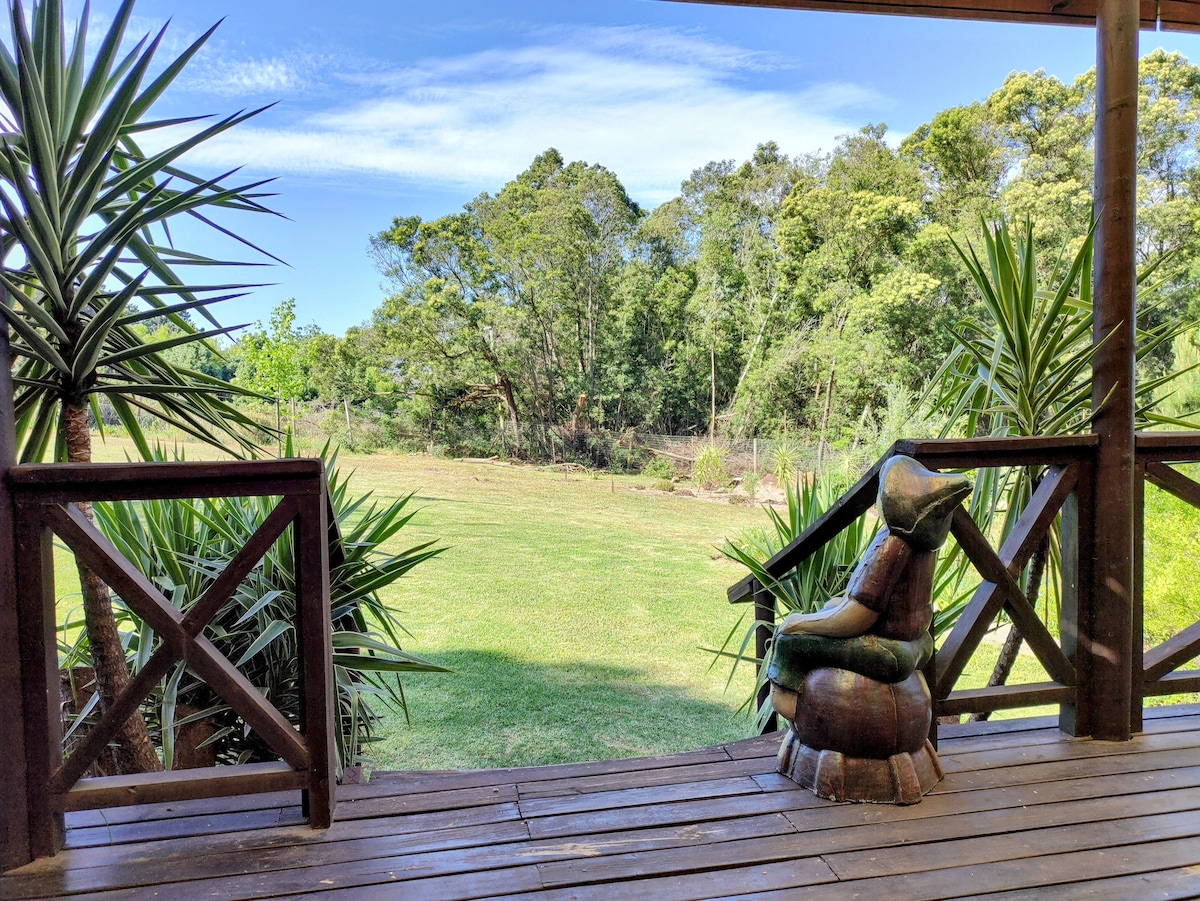 A wooden deck offers views of the expansive green lawn, surrounded by lush trees and shrubs. A carved wooden figure sits on the edge of the deck, inviting guests to relax and enjoy the serene natural surroundings.