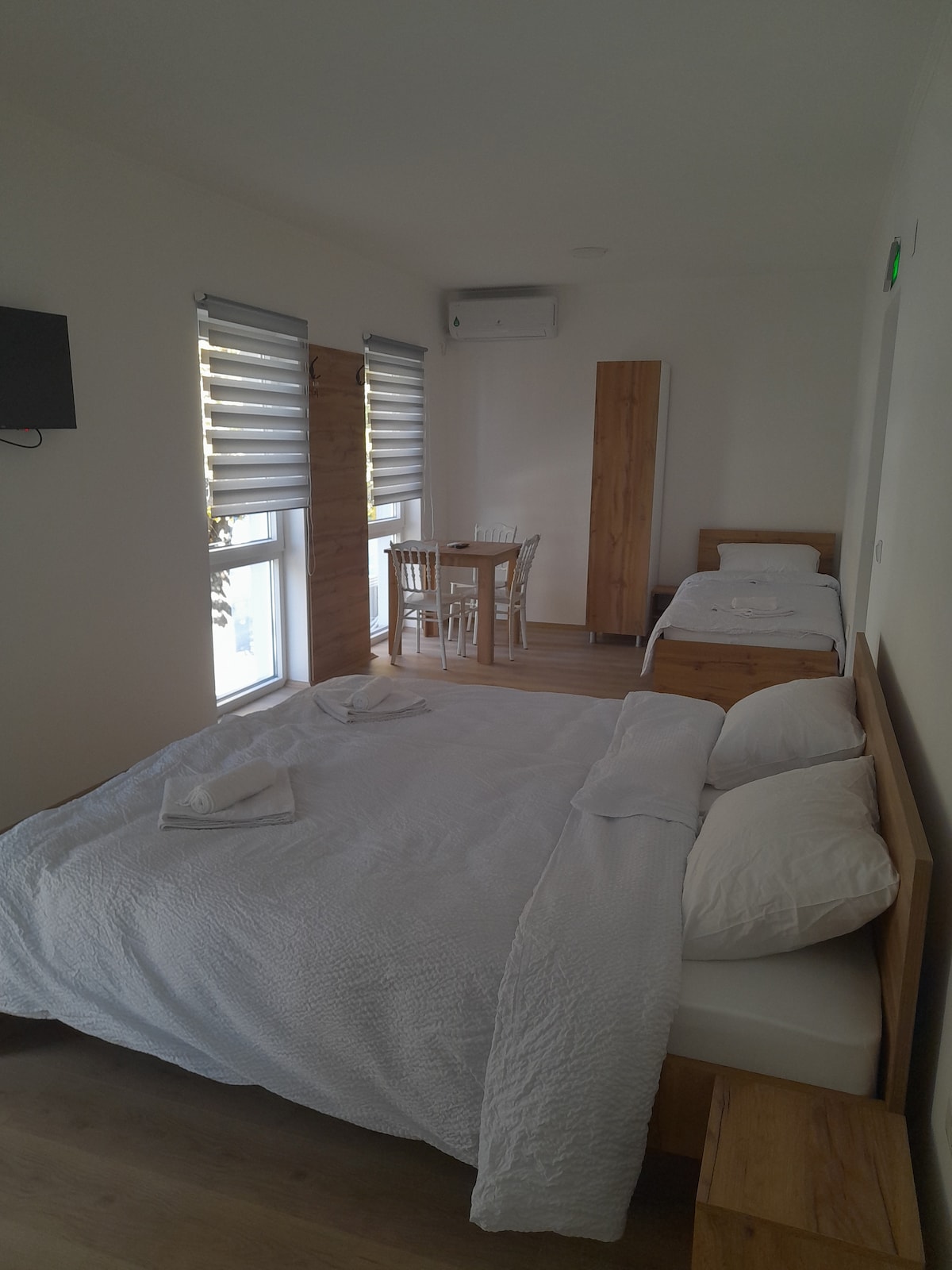 A well-lit room features a spacious bed with crisp white linens, paired with a separate single bed. A wooden dining table with chairs is positioned nearby. Two large windows are dressed with blinds, allowing natural light to filter in.
