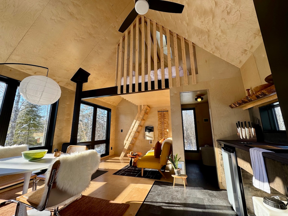 An open living area features high ceilings with wooden beams and large windows allowing natural light to fill the space. A cozy yellow armchair complements a dining table, while a kitchenette is neatly arranged along one wall. The upper loft is visible, showcasing additional sleeping space.