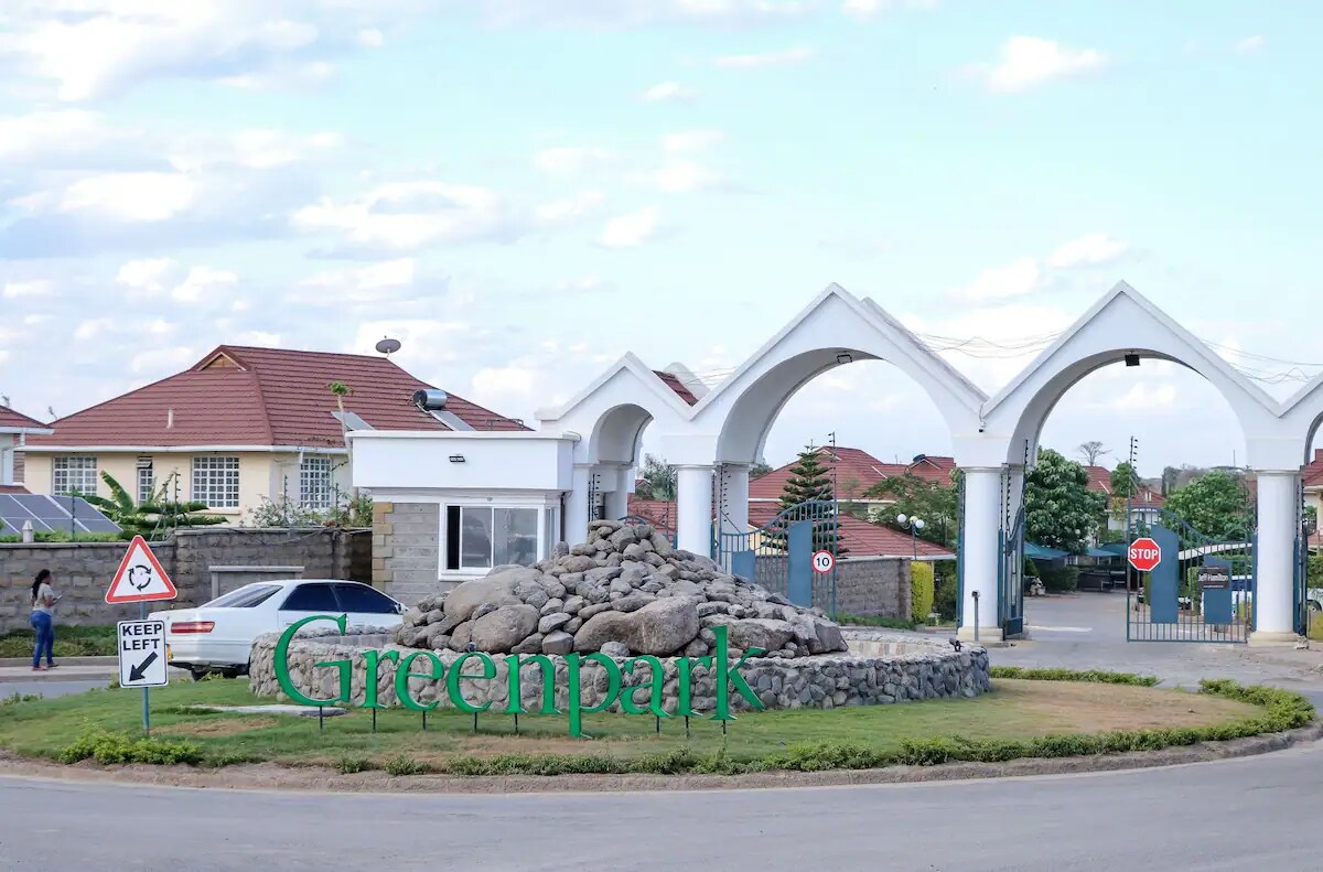 An entrance sign to the Greenpark neighborhood is displayed prominently at the roundabout, surrounded by a rock formation. The archway structures are visible, leading into the residential area, while vehicles are seen parked by the roadside and traffic signs indicate driving directions.