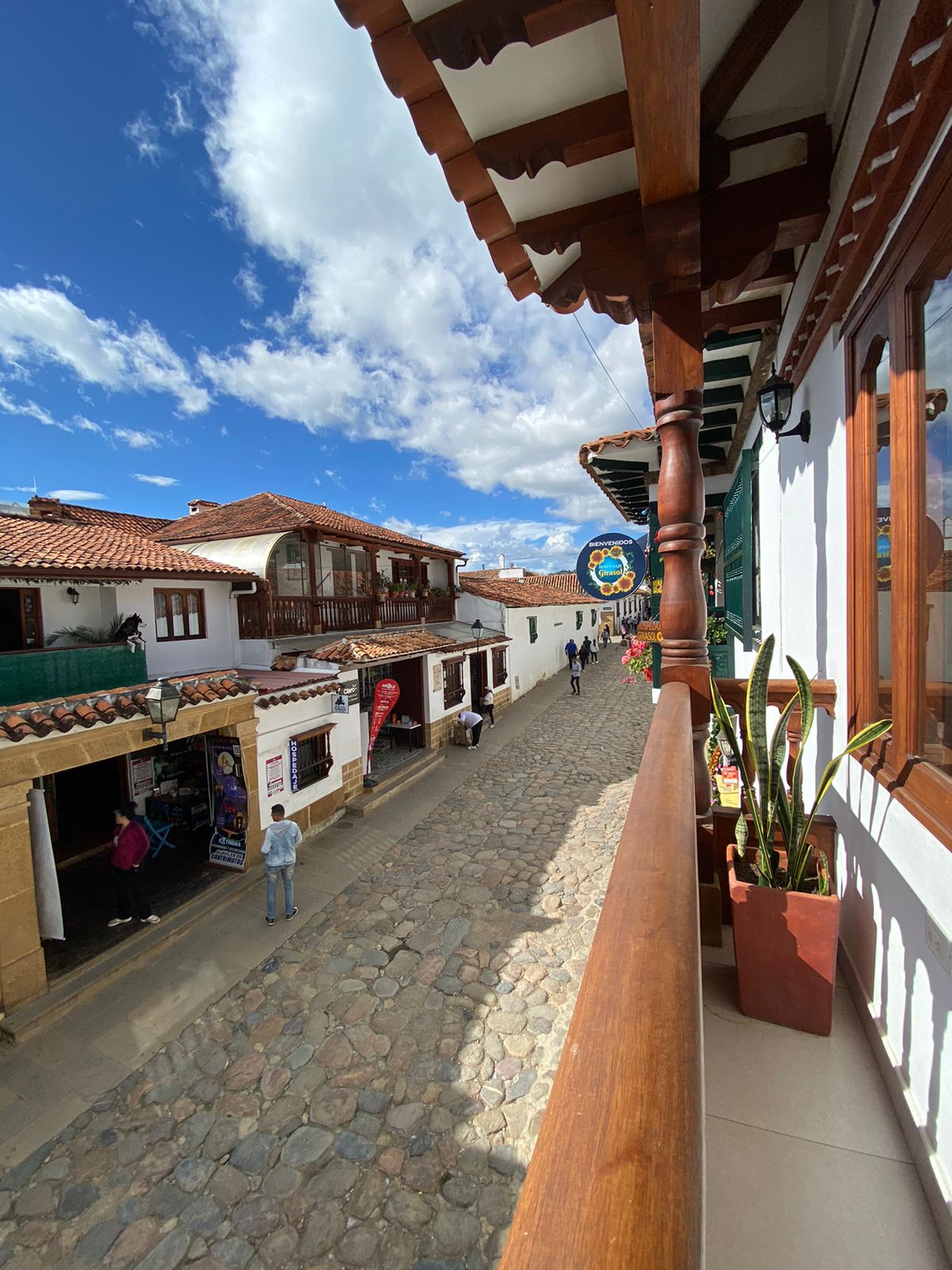 A cobblestone street is framed by traditional buildings featuring wooden balconies and colorful facades. The sky is partially cloudy, providing a bright atmosphere. Potted plants adorn the balcony, adding a touch of greenery to the view of the bustling street below.