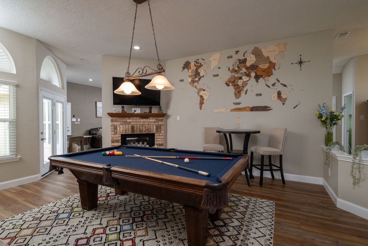 A spacious game room is highlighted by a classic pool table, its green felt contrasting with rich wooden accents. A large map made of varied materials adorns the wall, while soft lighting from overhead fixtures illuminates the area. A seating area includes two high-backed chairs next to a small table.