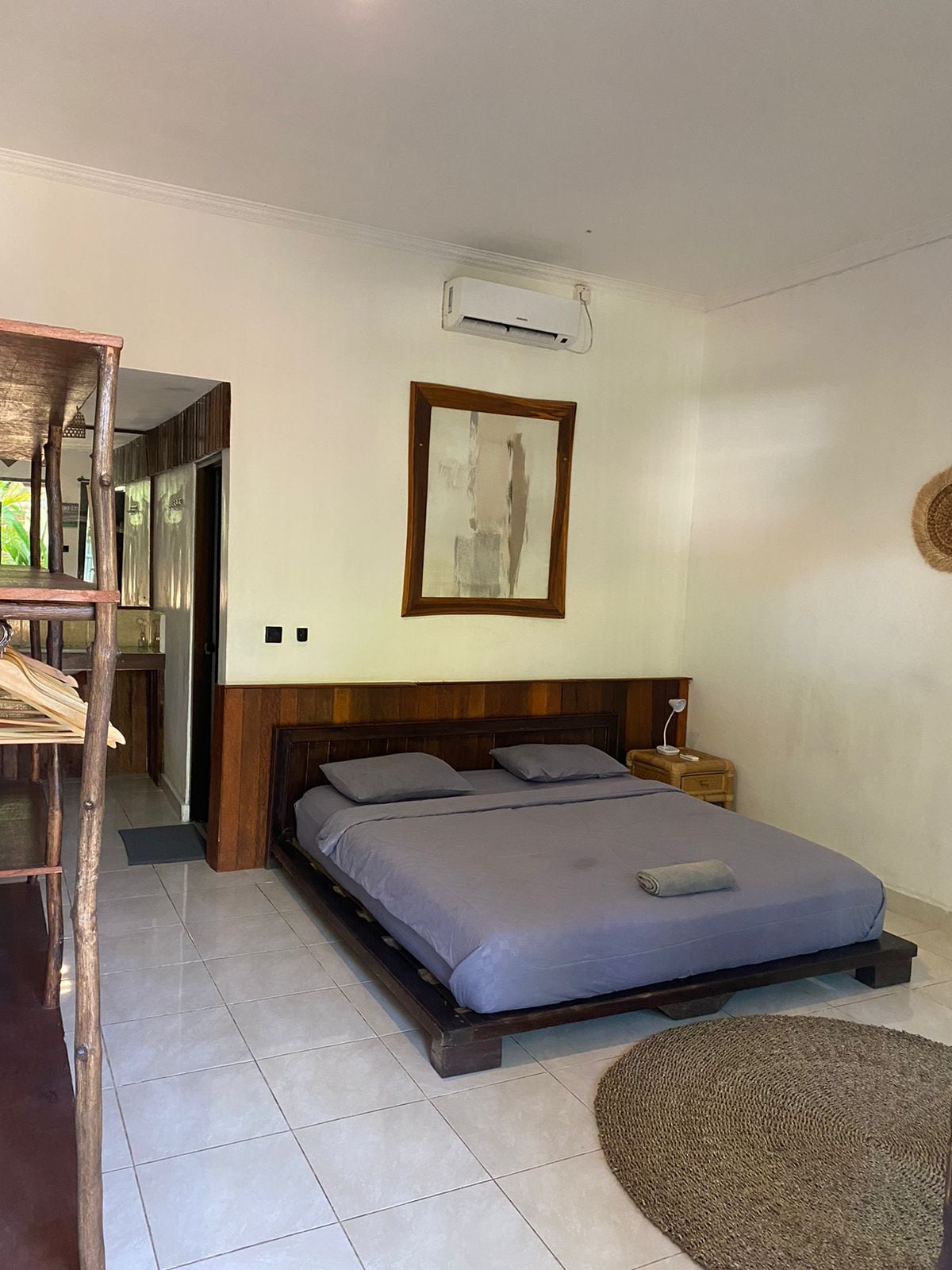 A comfortable sleeping area features a spacious bed with a neutral-toned bedding set, complemented by a woven rug beneath. A framed artwork hangs on the wall, while a small lamp and a wicker chair add functionality. Air conditioning is visible above the bed.