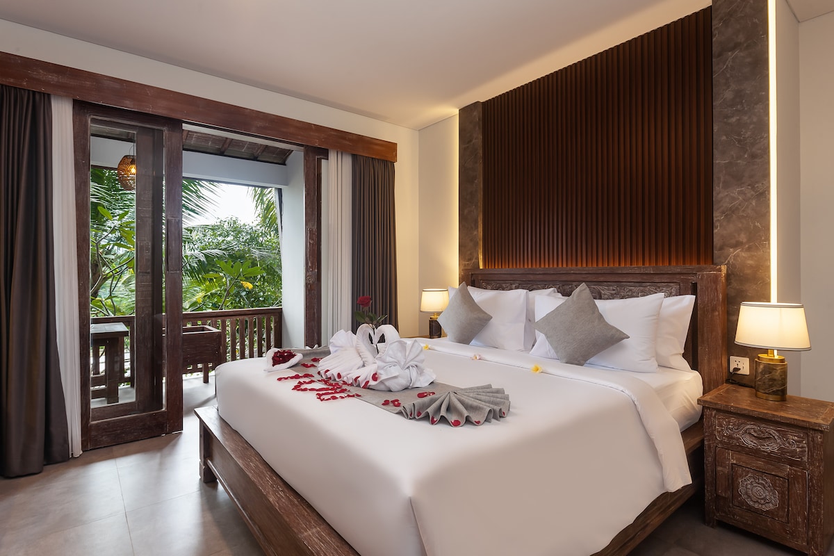 A spacious bedroom is adorned with elegant wooden accents and soft lighting. The bed is neatly made with white linens, complemented by decorative gray pillows. Double doors open to a balcony surrounded by lush greenery, inviting natural light into the room.
