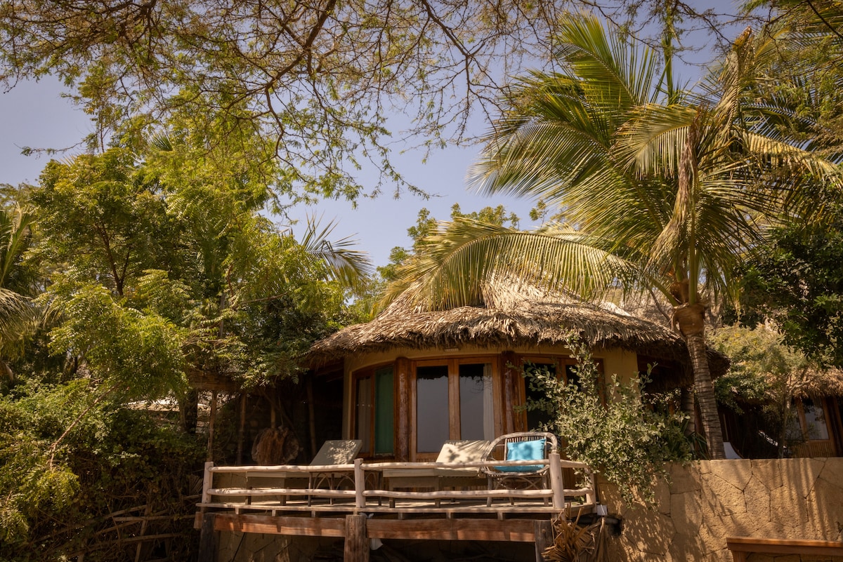 A rustic bungalow features a thatched roof surrounded by lush greenery and palm trees. A private balcony is visible, with outdoor seating overlooking the natural surroundings. Large windows allow ample light into the interior, creating a connection with the outdoors.