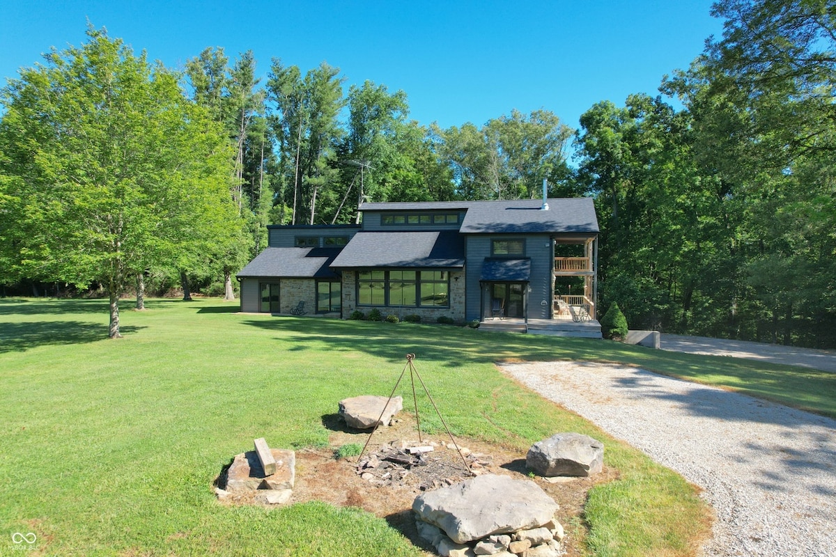 A modern wooden cabin is set amidst expansive green lawns, bordered by trees. The structure features large windows, allowing for natural light. A fire pit, constructed of stones, is situated in the foreground, surrounded by grass, inviting gatherings under the open sky.