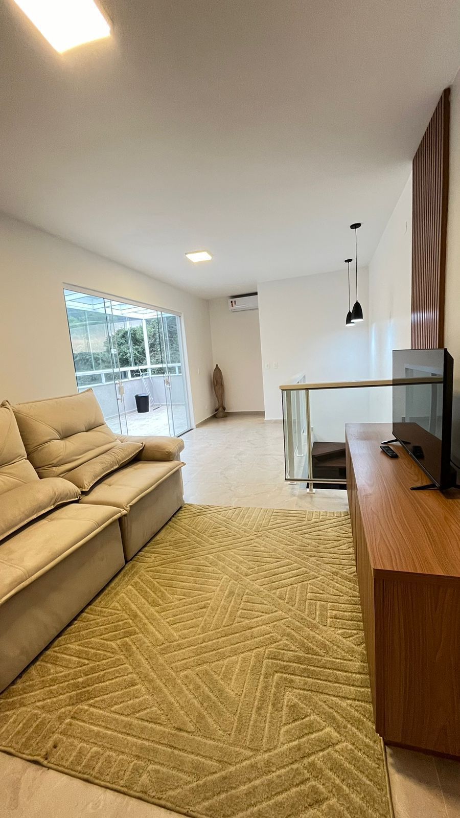 A spacious living area is furnished with a light-colored sofa facing a wooden media console. A large glass sliding door allows natural light to fill the space while providing access to an outdoor balcony. A soft area rug adds comfort beneath.
