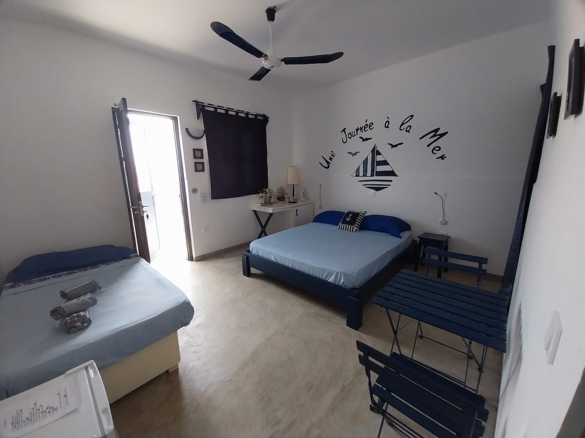 A bright bedroom features a spacious double bed and a separate single bed, both adorned with blue linens. A small blue table and chairs are positioned near the window, which is partially covered by dark curtains, allowing natural light to enter the room.
