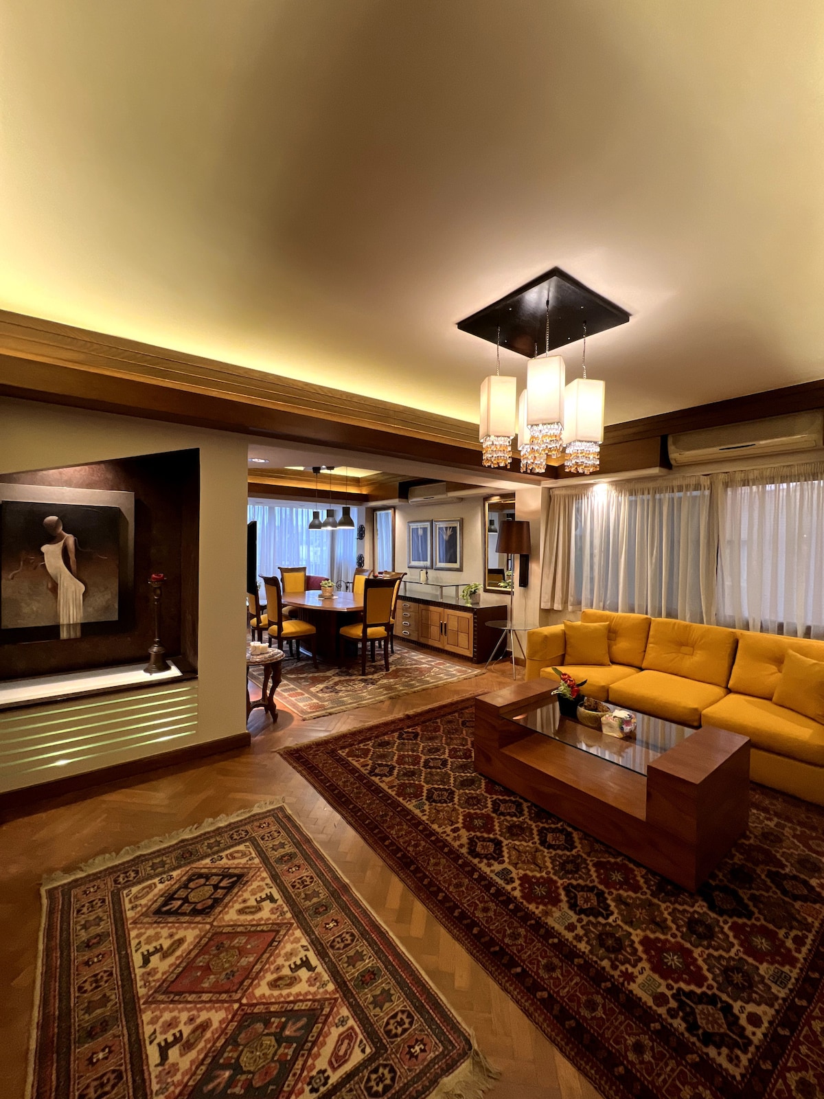 A spacious living area features a warm yellow couch and a wooden coffee table, complemented by an ornate area rug. Nearby, a dining area showcases a wooden table surrounded by matching chairs, while soft lighting highlights the artwork on the wall.