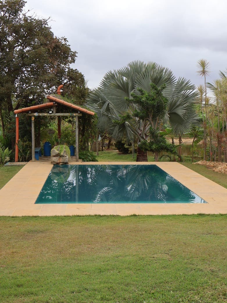 A well-maintained swimming pool is surrounded by lush greenery, featuring a shaded wooden structure nearby. The pool reflects the overcast sky, while palm trees and various plants enhance the natural setting.