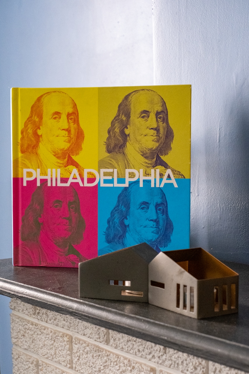 A colorful book titled 'Philadelphia' is displayed with a vibrant cover featuring multiple found portraits, with facial features obscured. Next to the book sits a small metal house-shaped sculpture, resting on a dark stone surface against a light blue wall.