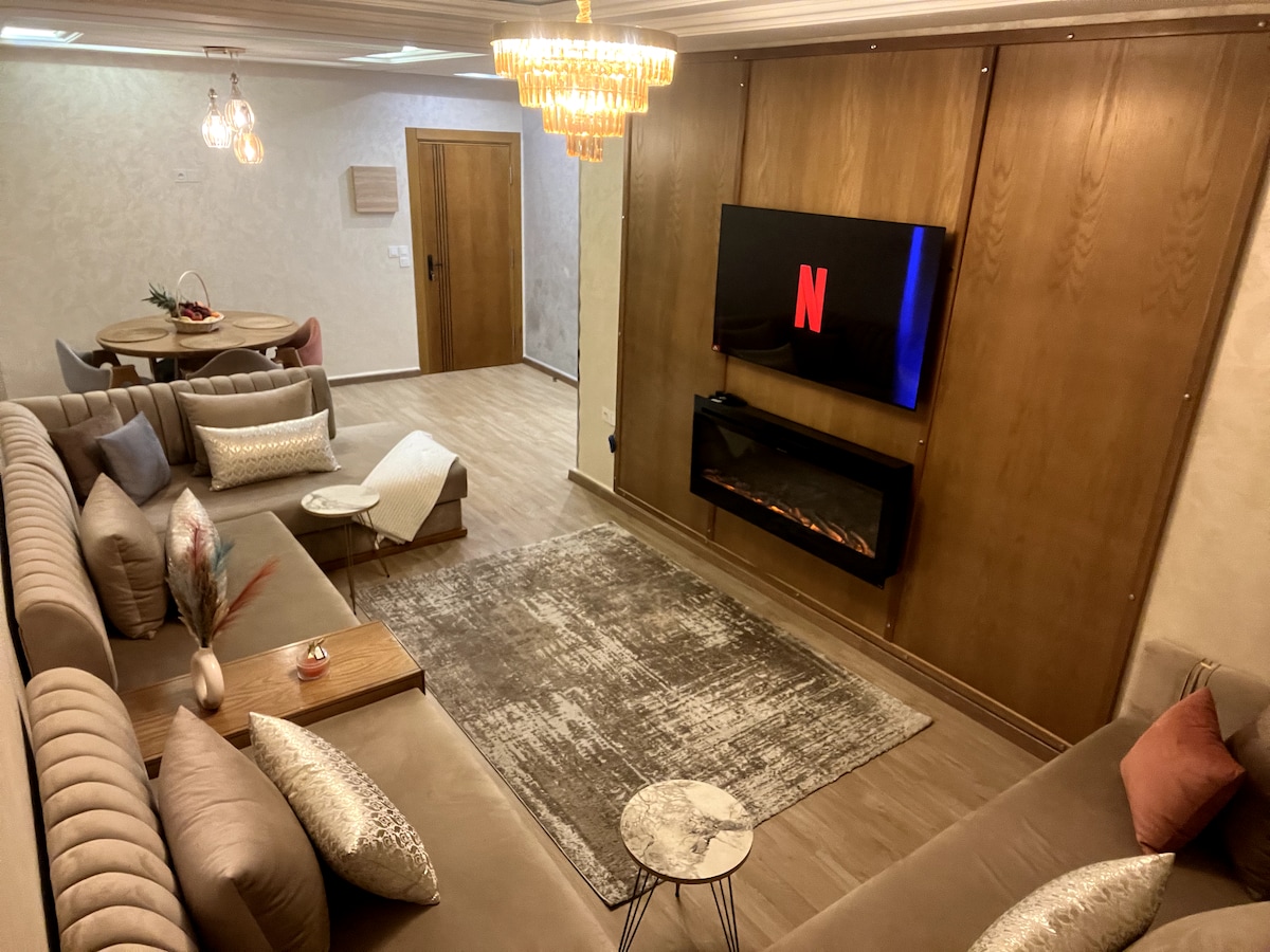 The living area is showcased with comfortable seating arranged around a modern coffee table. An electric fireplace adds a touch of warmth, while a large TV mounted on the wall provides entertainment options. Soft lighting is created by a stylish chandelier above.