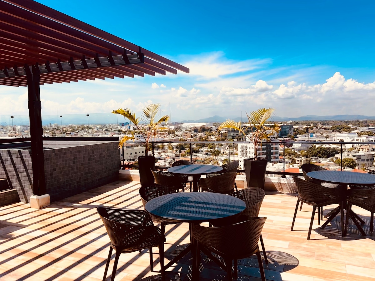 An outdoor terrace area is presented with several round tables and black chairs. Potted plants are placed strategically, adding green accents. The skyline of Santo Domingo is visible in the background, with a clear blue sky and distant mountains enhancing the scenic view.