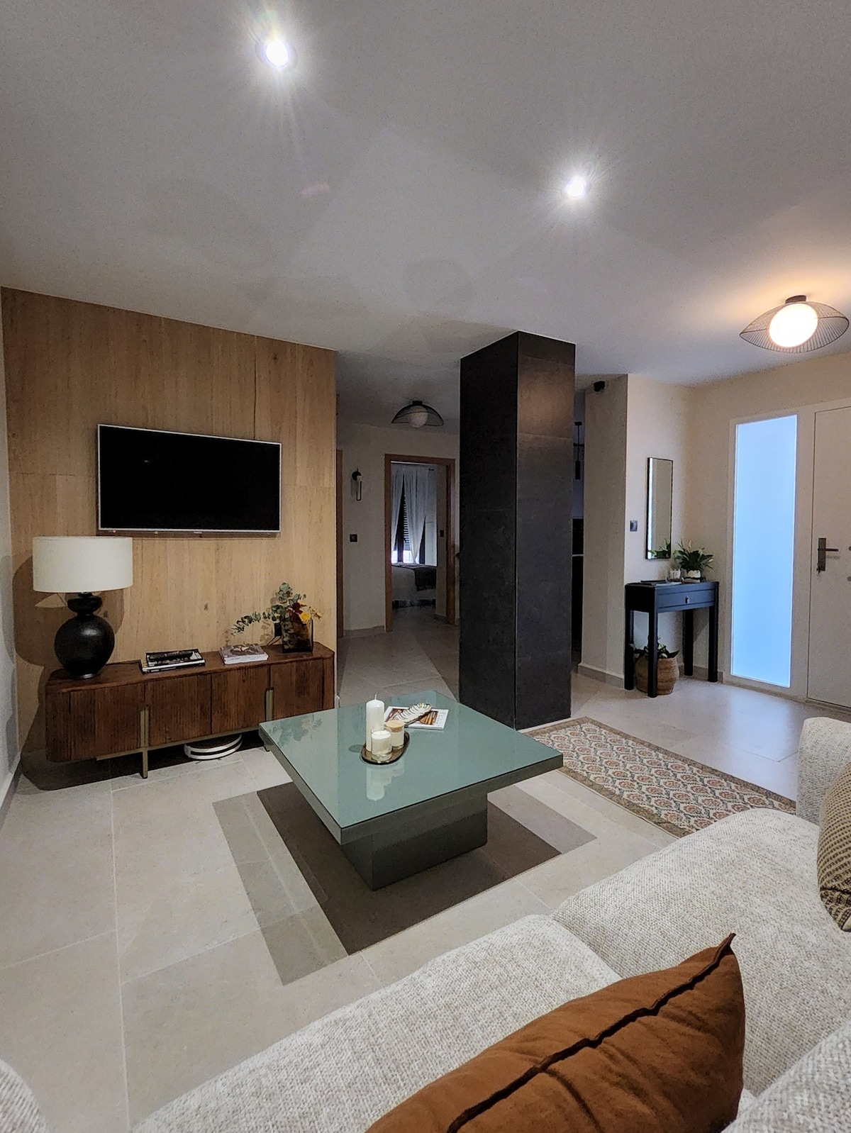 A modern living area is presented with a wall-mounted television above a wooden media console. A glass coffee table is centered on a light tile floor, surrounded by a soft sofa. Soft lighting highlights the welcoming ambiance of the space, leading towards an adjoining hallway.