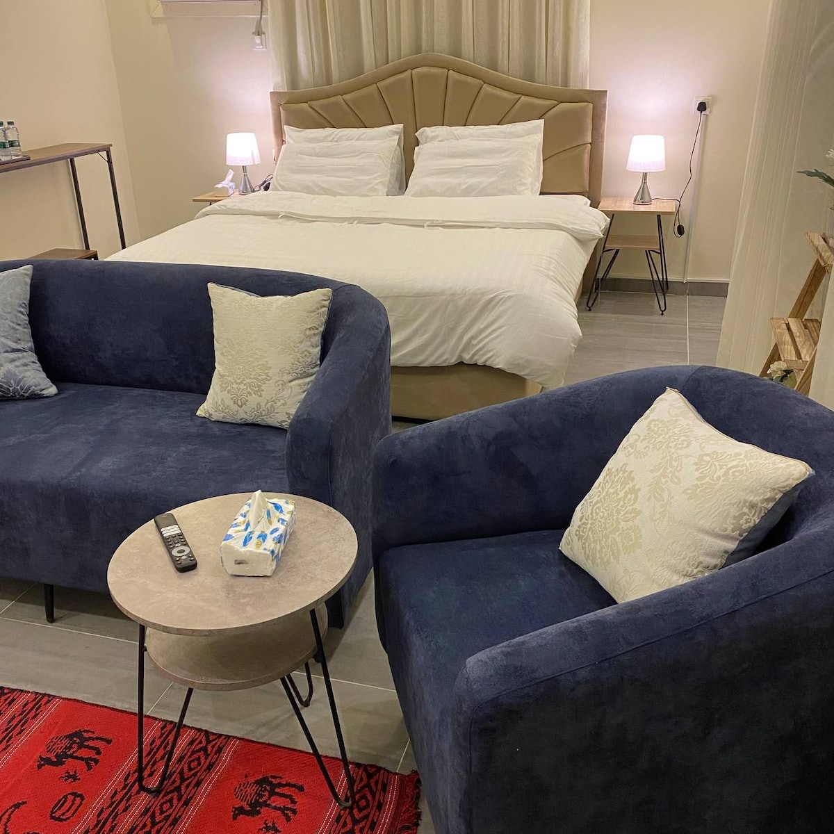 A cozy sleeping area features a spacious bed with neatly arranged linens. Two plush chairs with soft cushions are positioned near a small table, which holds a remote control and a decorative item. Soft lighting is provided by lamps on either side of the bed.