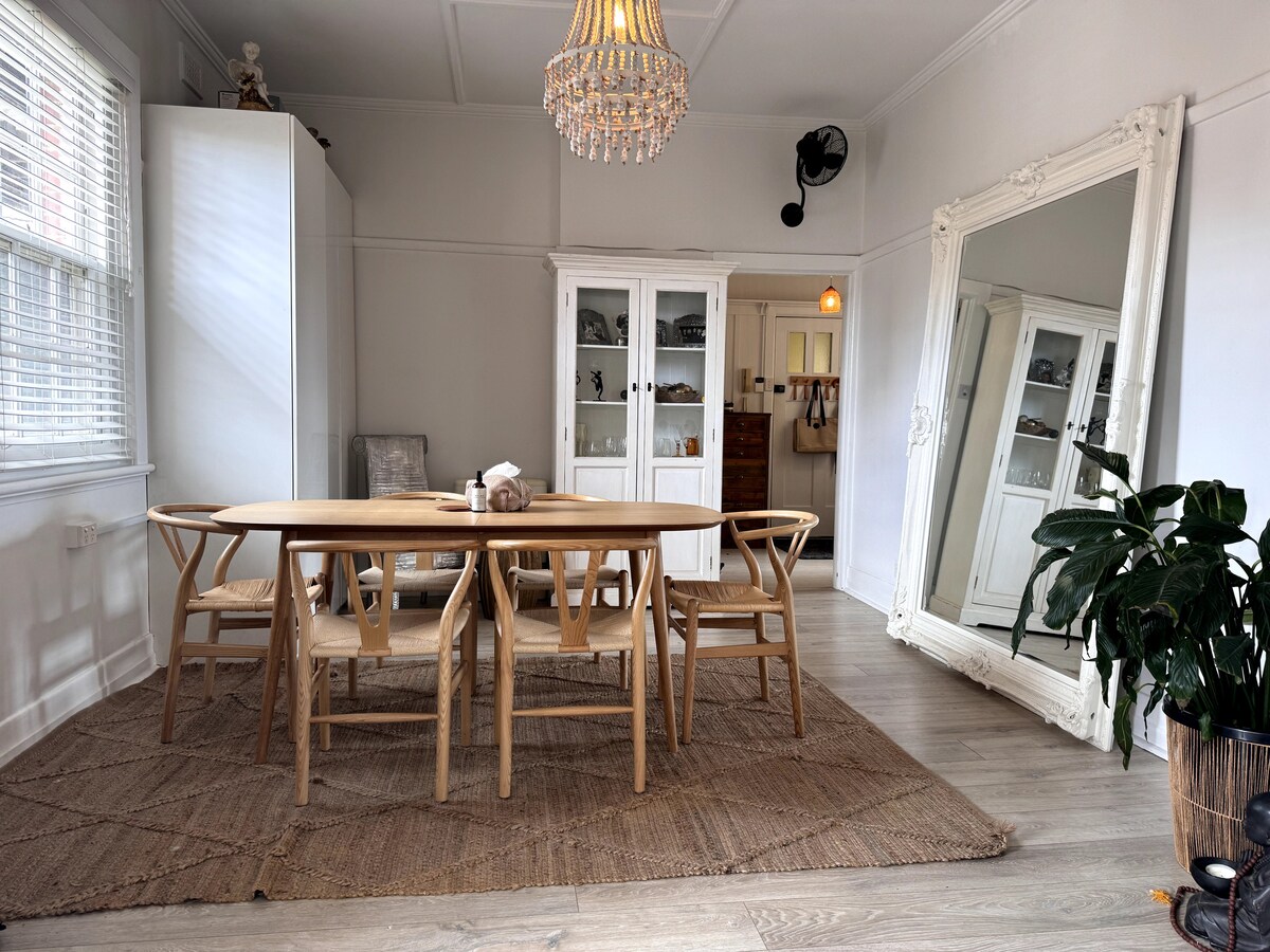 A light-filled dining area features a wooden oval table surrounded by six chairs, set on a textured beige rug. A tall, vintage mirror reflects the space, while a tall plant adds a touch of greenery. Large windows bring in natural light, enhancing the airy feel.