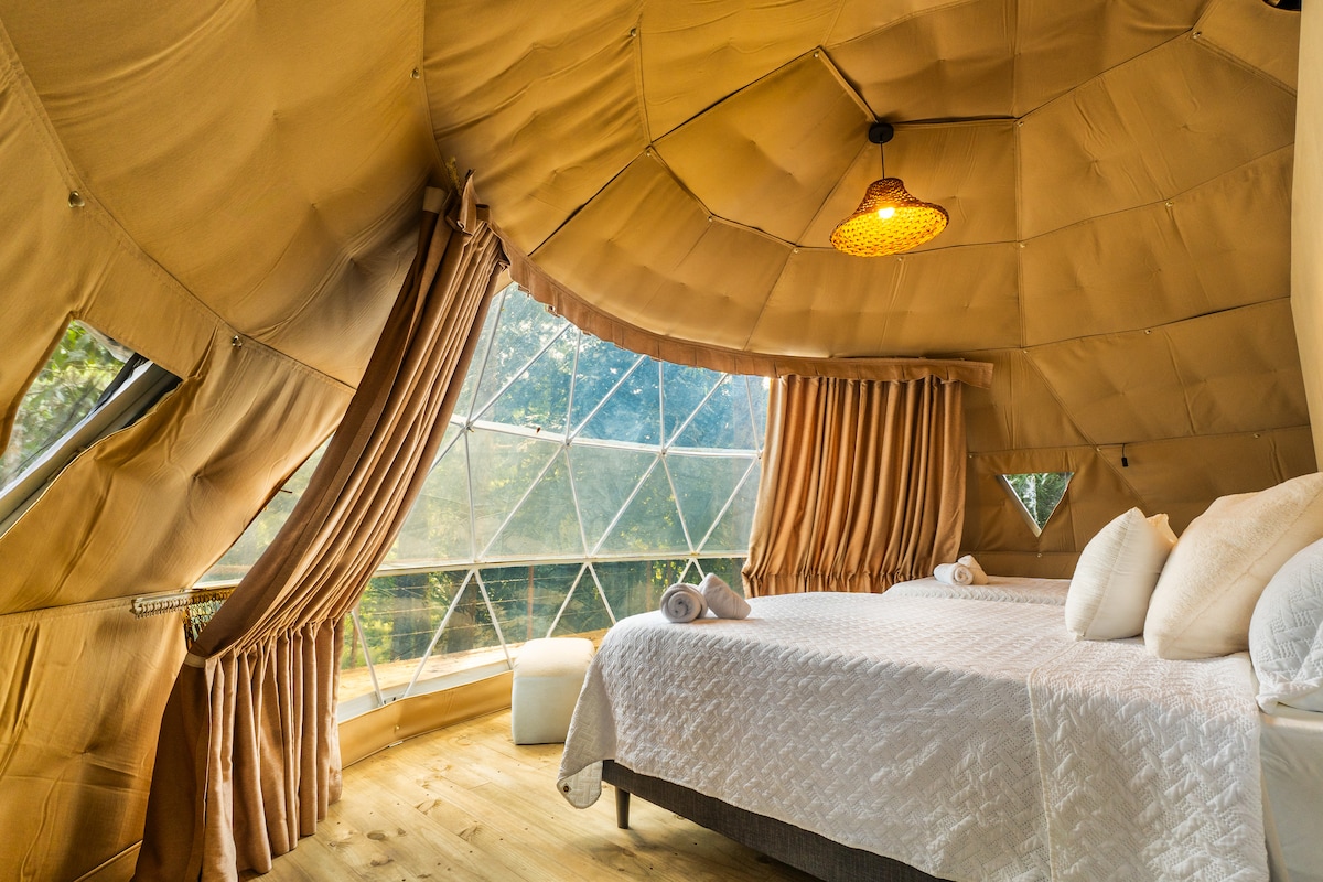 The cozy sleeping area features a spacious bed adorned with light bedding and decorative pillows. Large windows allow for abundant natural light and provide views of the surrounding landscape. Soft curtains frame the windows, complementing the warm tones of the interior.