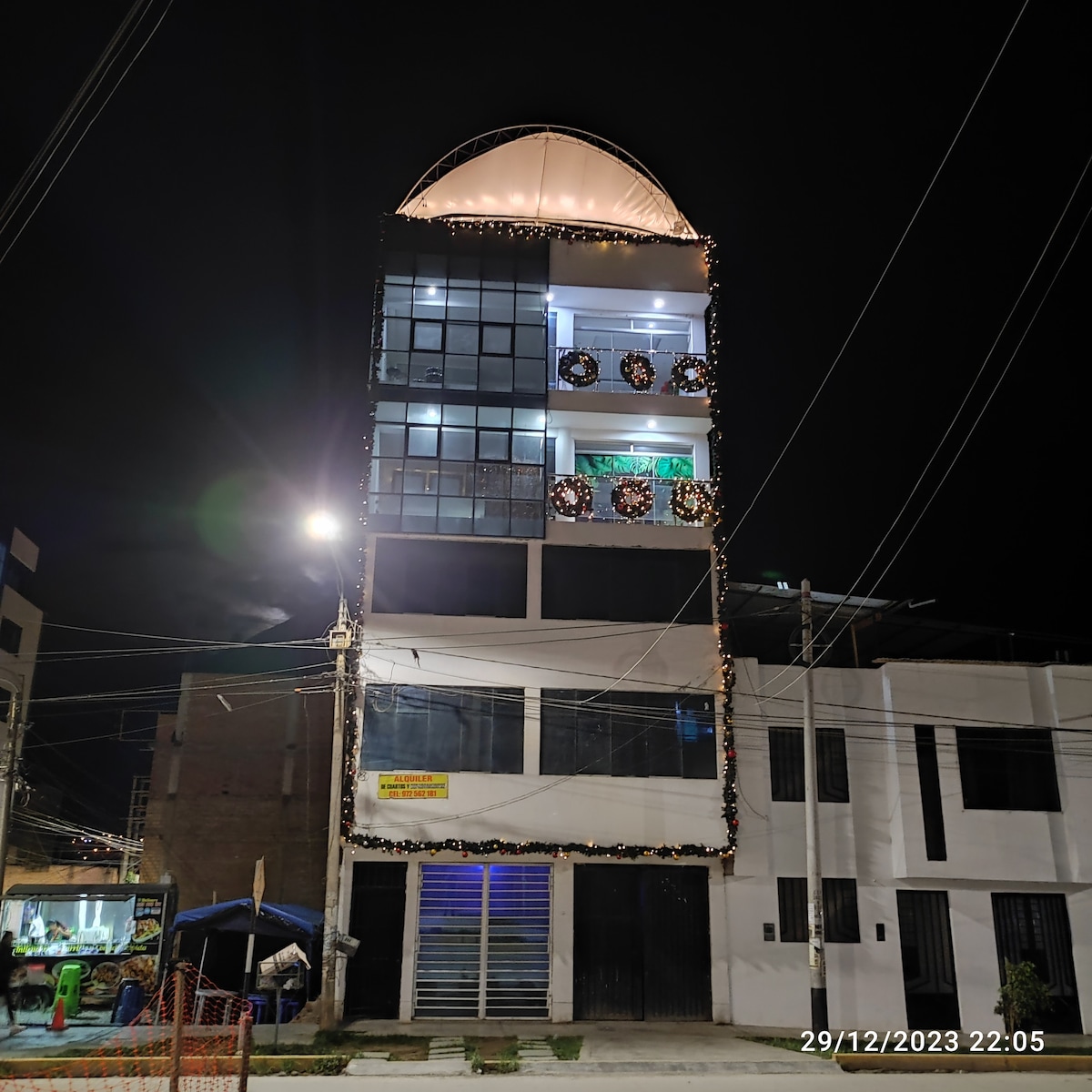 The exterior of a multi-story building is adorned with festive decorations. Its modern design features large windows and a distinctive arched roof. Lights illuminate the façade against the night sky, highlighting the holiday wreaths placed symmetrically along the upper levels.