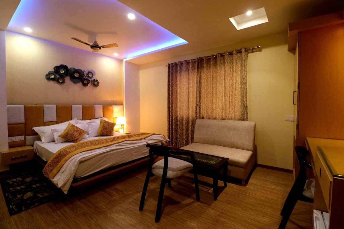 An elegant room features a plush bed with decorative pillows, complemented by a cozy seating area. Soft lighting from the ceiling and wall art enhances the ambiance. A wooden desk and chair provide a workspace, while curtains frame the window, allowing natural light to filter in.